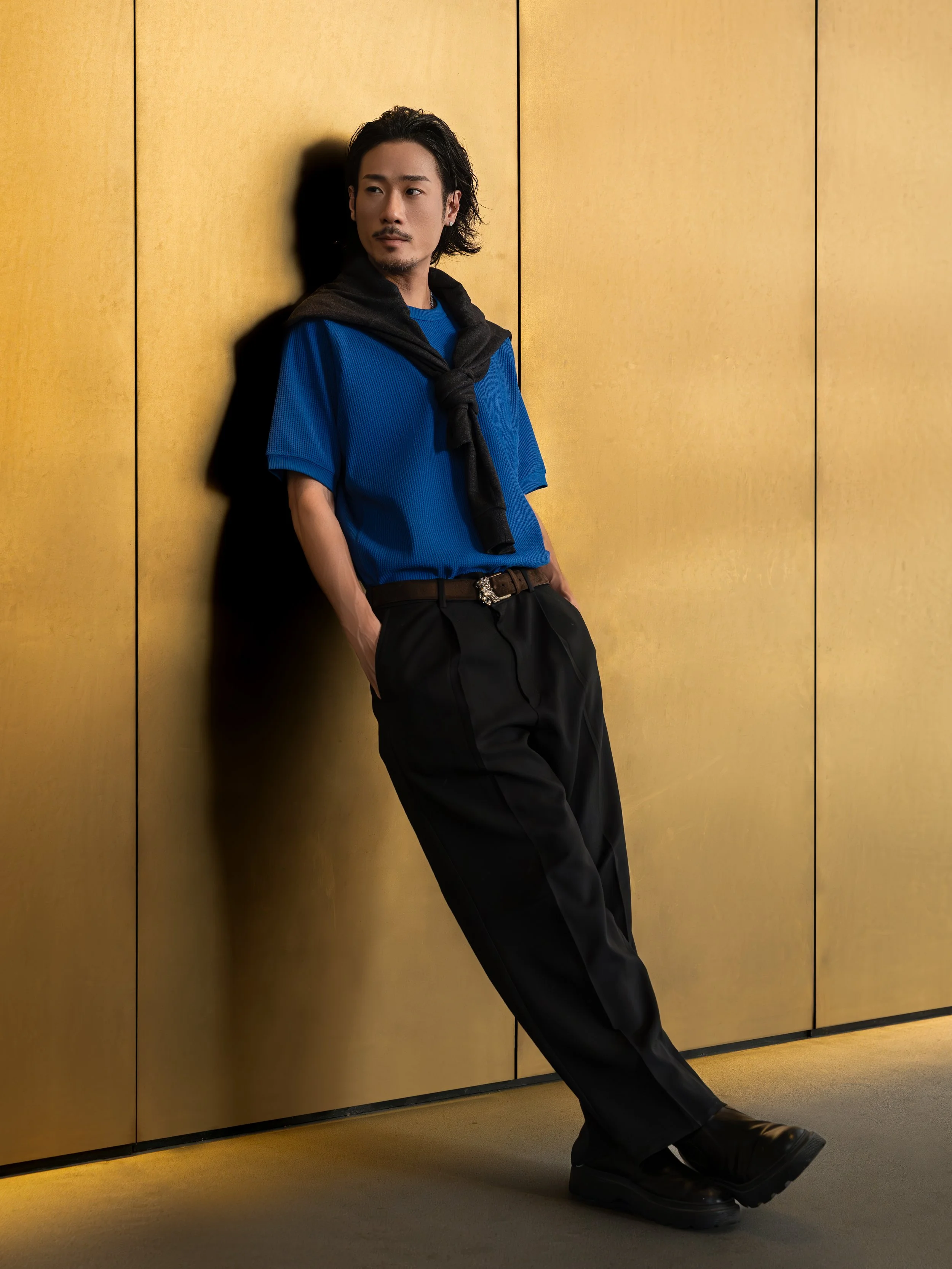 A man with long dark hair, facial hair, wearing a blue shirt, black pants, and boots, leaning against a golden wall, with a black sweater draped over his shoulders.