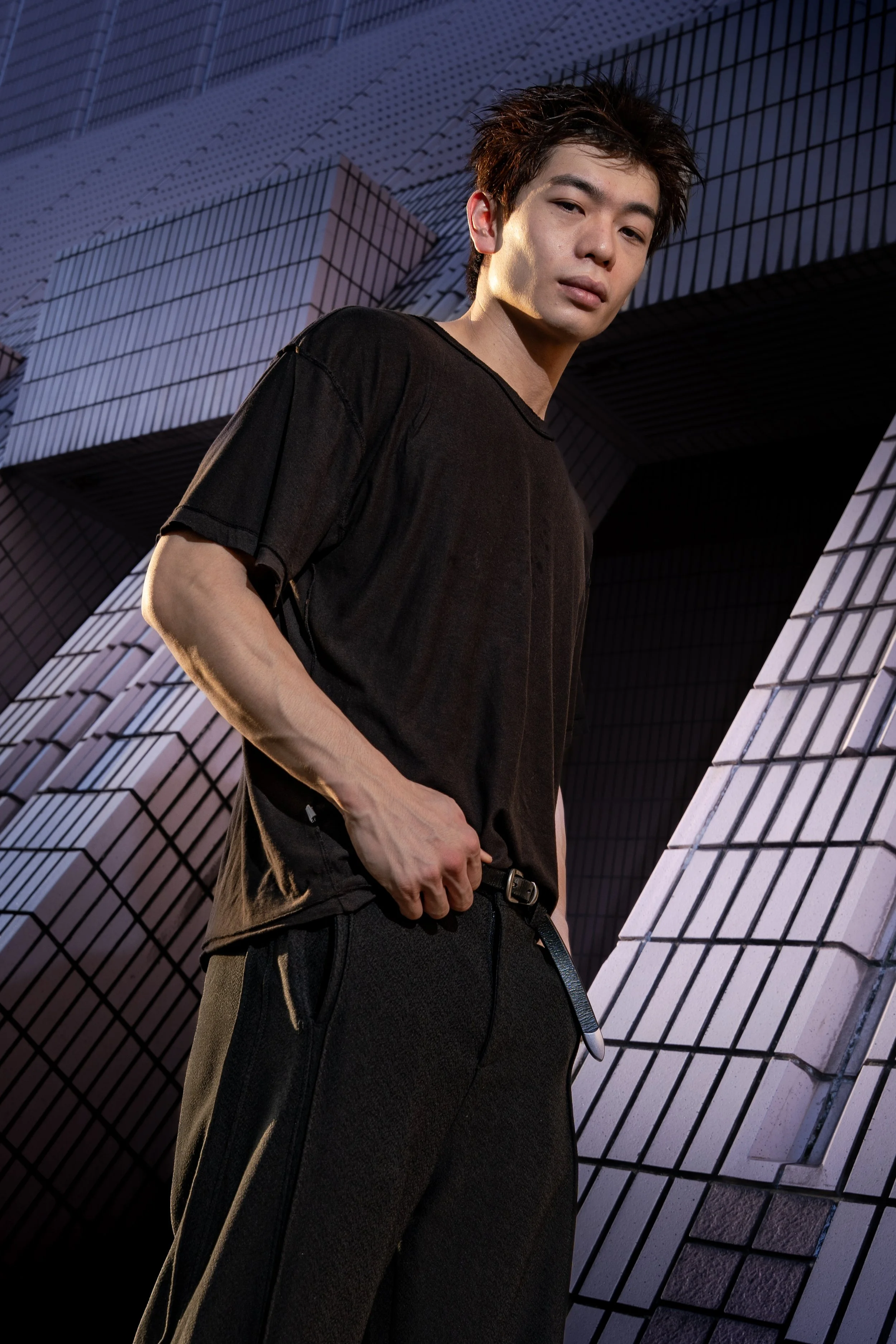 A young man with dark hair and light skin wearing a black t-shirt and black pants standing in front of a modern building with a geometric, tiled design.