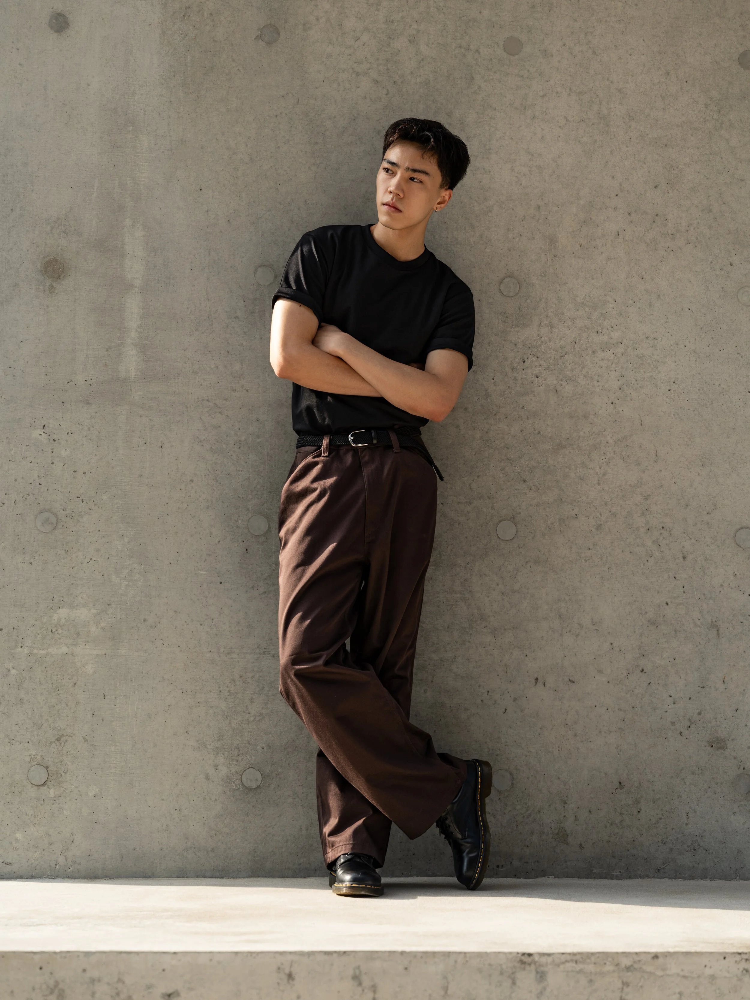 Young man leaning against a concrete wall with arms crossed and looking to the side, wearing a black t-shirt, brown pants, and black shoes.