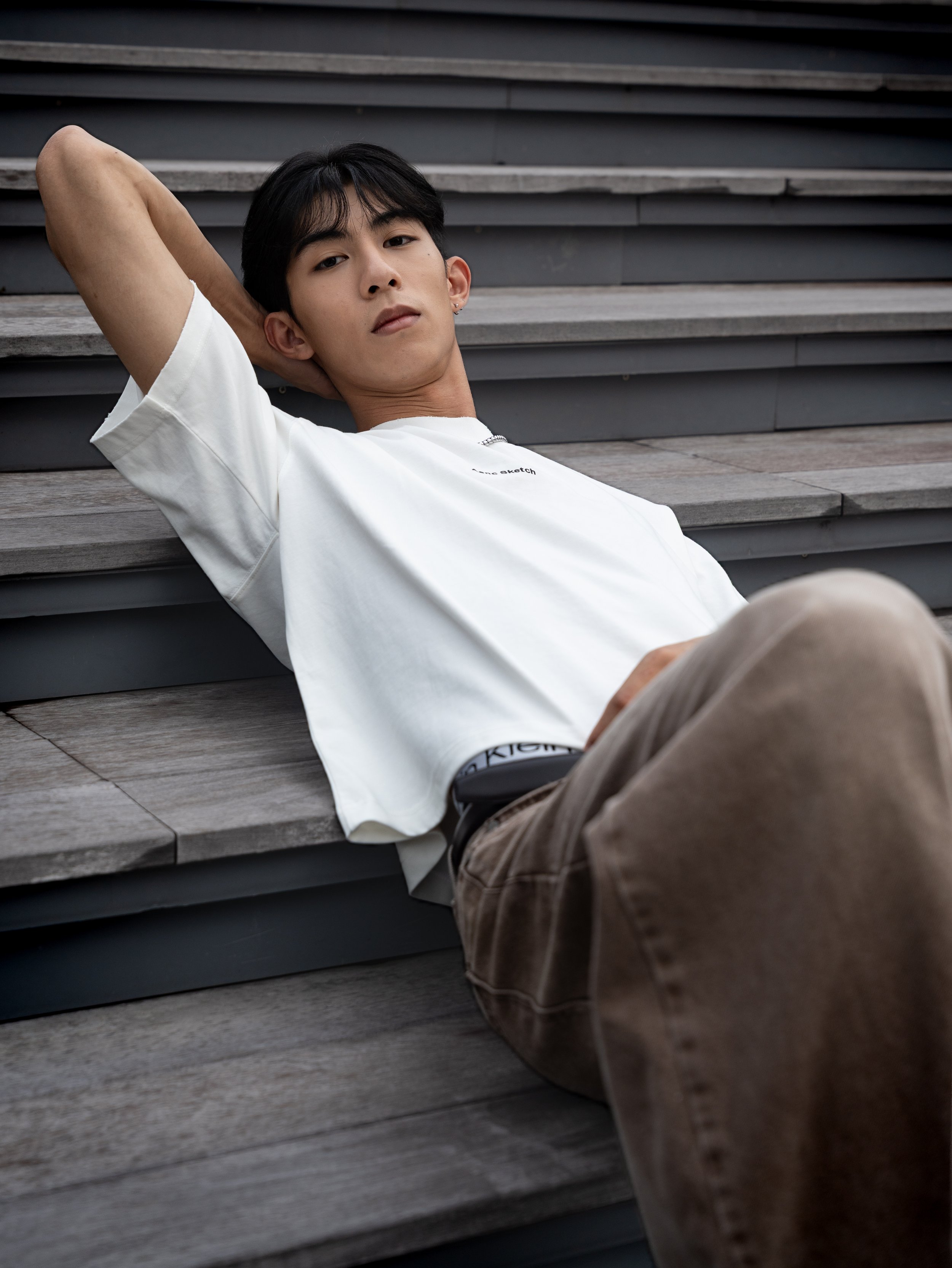 A young man with black hair reclining on wooden stairs, wearing a white t-shirt and brown pants, looking at the camera with a calm expression.