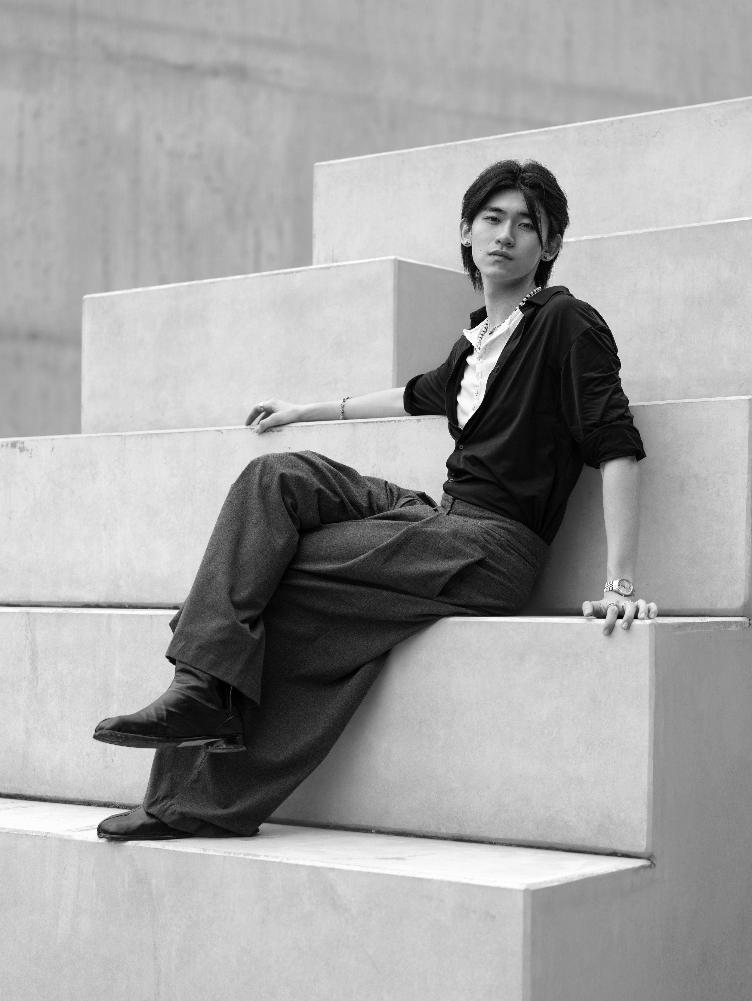 A young man sitting on steps outside, wearing a dark shirt and pants, with one arm resting on the step and the other hand on his knee, looking at the camera. Black and white photograph.