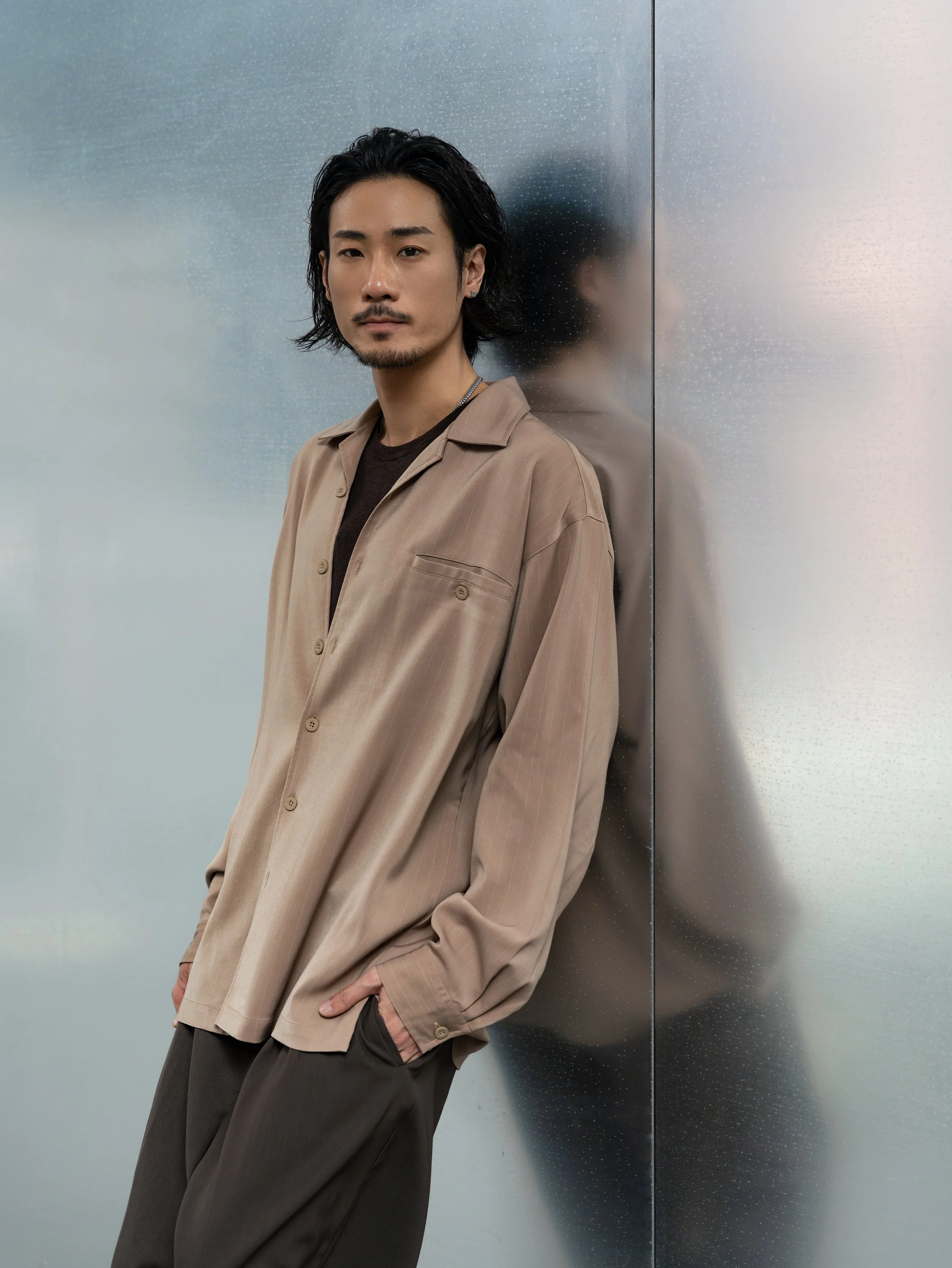 A man with black hair, a mustache, and a goatee stands against a speckled metallic surface. He is wearing a beige jacket over a dark shirt, with hands in his pockets, looking at the camera.