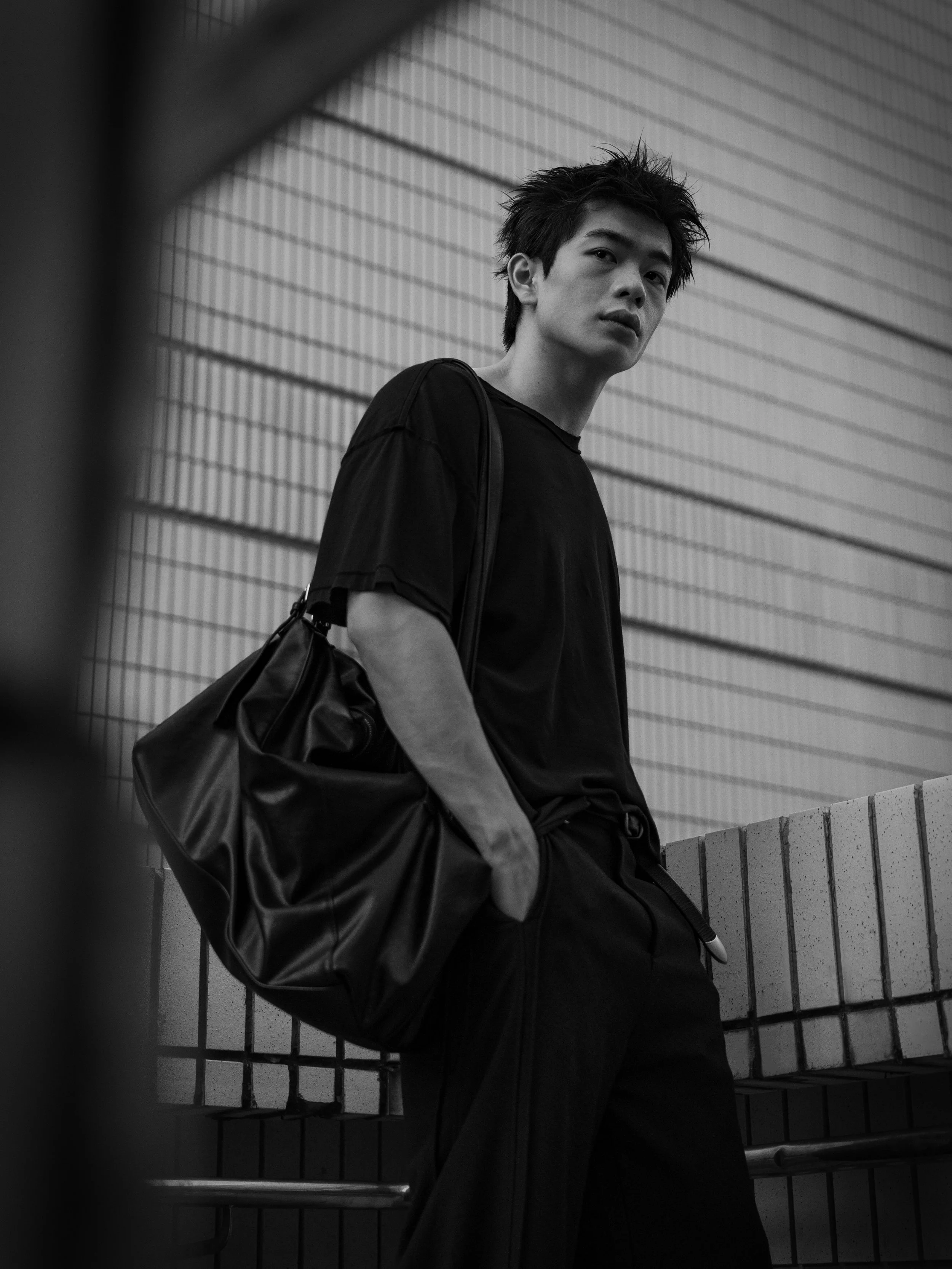 A young man with spiky hair wearing a black t-shirt and carrying a black bag, standing against a tiled wall in an urban setting, with a metal railing in the foreground.