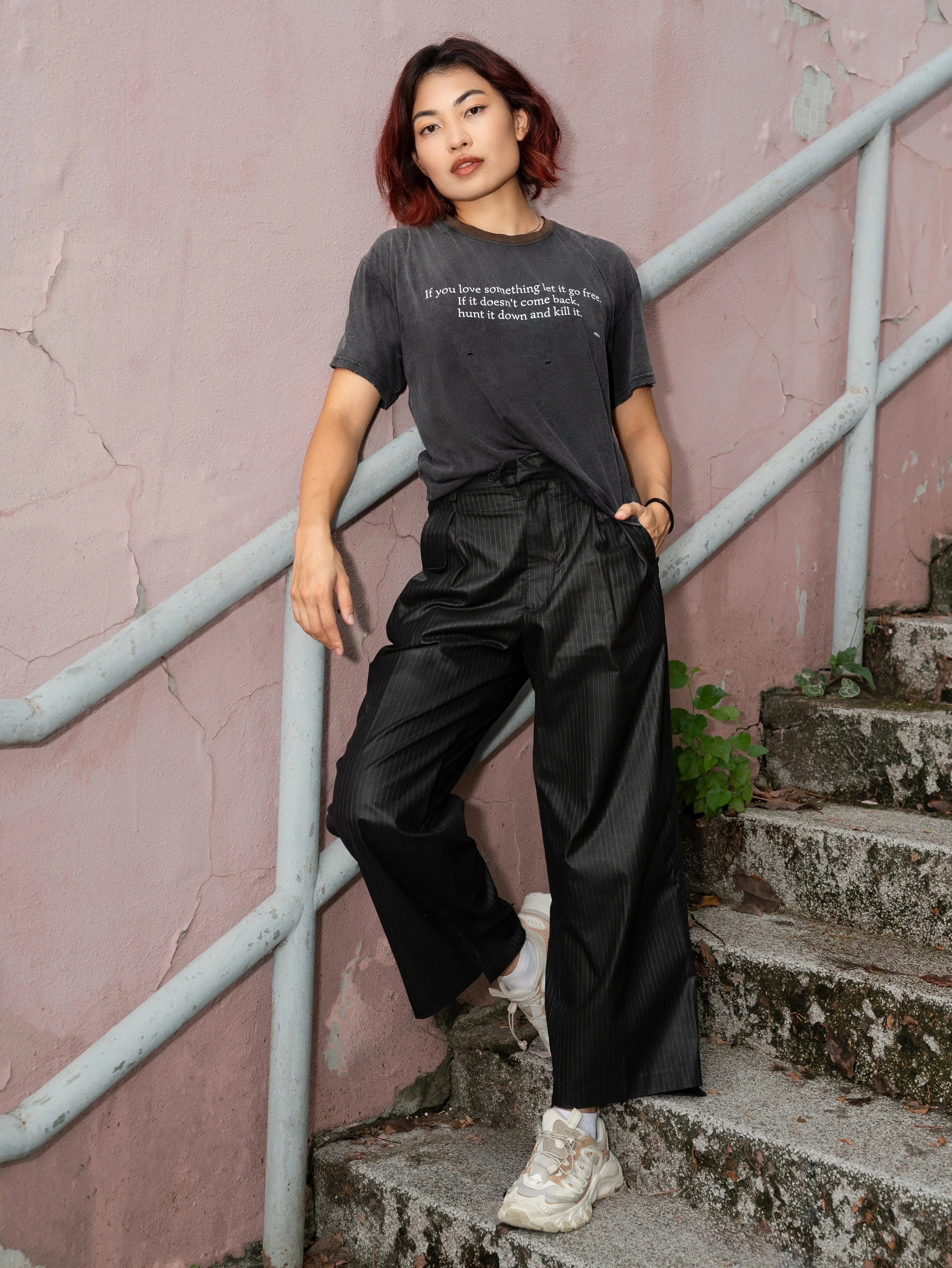 A young woman with shoulder-length, wavy, reddish-brown hair, standing on outdoor steps with a pink, cracked wall behind her. She is wearing a black graphic t-shirt, black pinstripe wide-leg pants, and off-white sneakers, looking confidently at the c