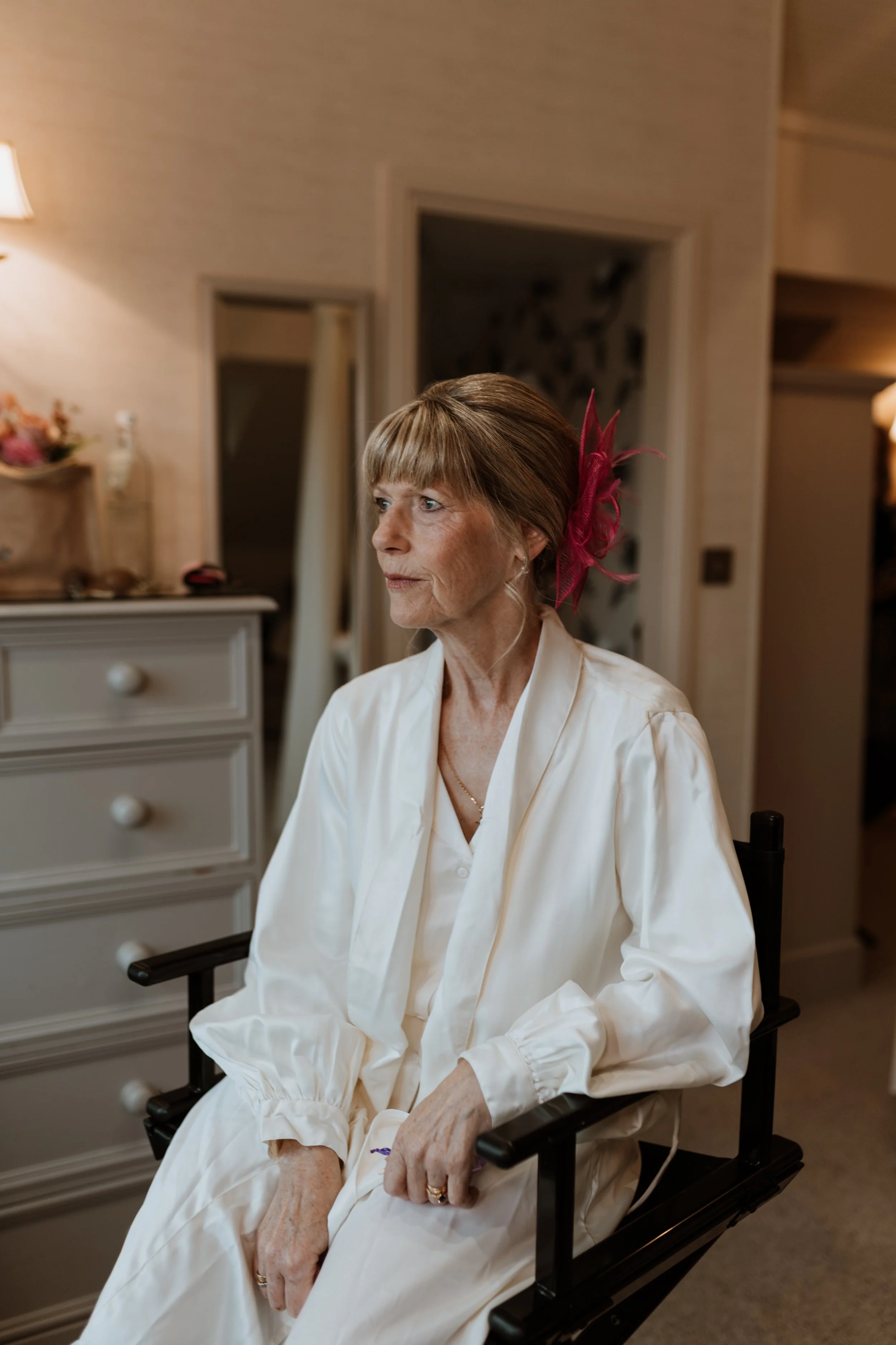  The mother of the bride is sits in a chair in a dressing gown as she gets her make up and hair done.  