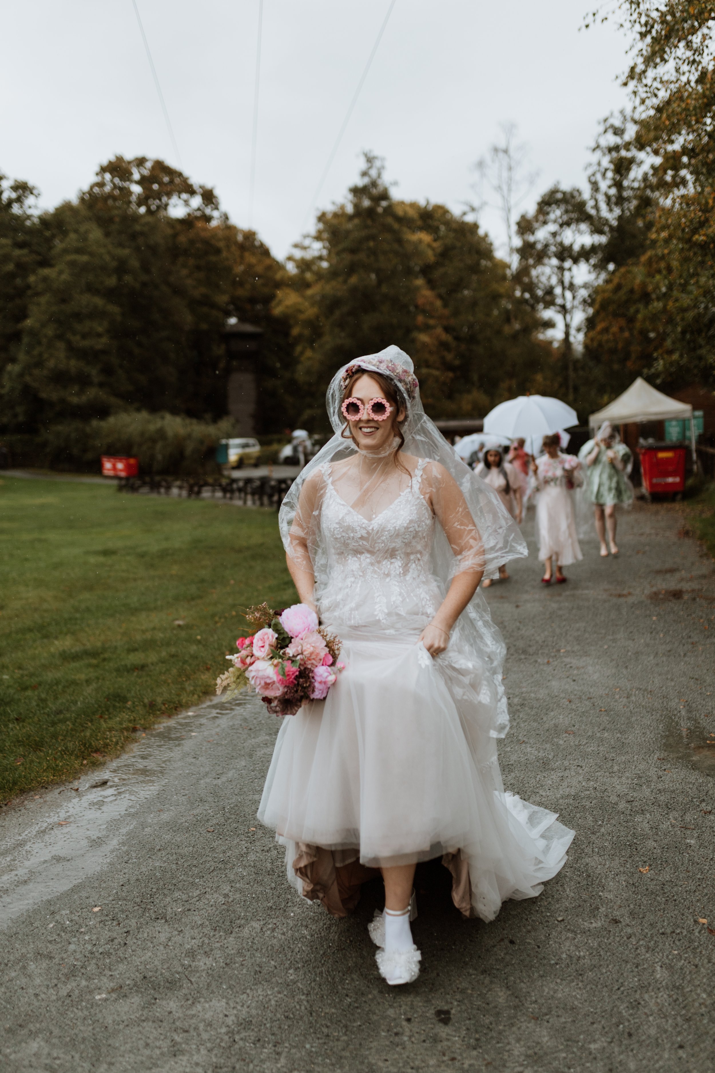  A bride wears a clear poncho as she walksin the rain in the Lake District, she has flower sunglasses on and is holding her flowers.  