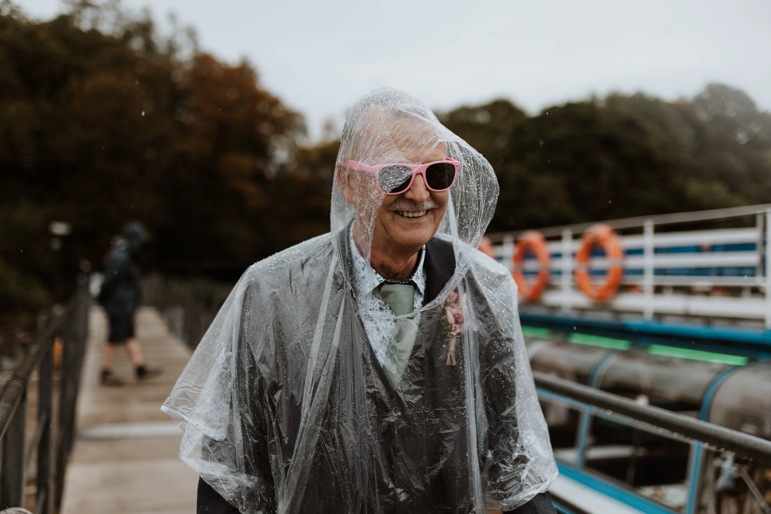  The brides father wears a poncho and flower sunglasses in the rain in the Lake District.  