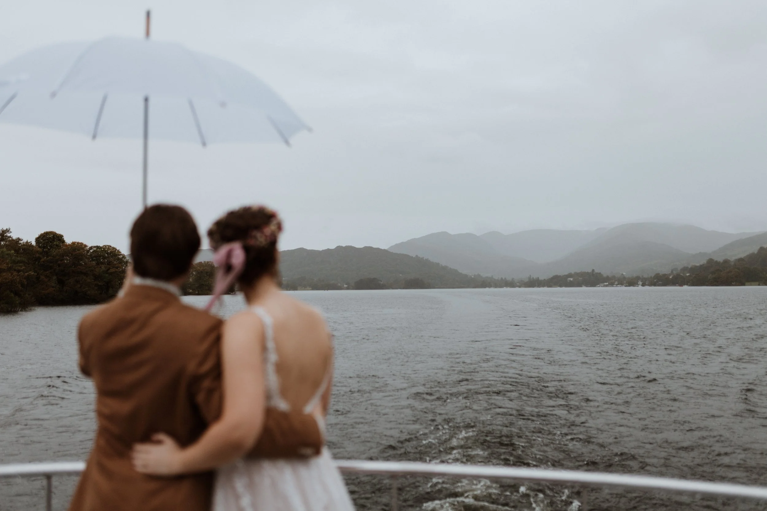  Bride and groom look out at the views under an umbrella during a rainy Lake District wedding. 