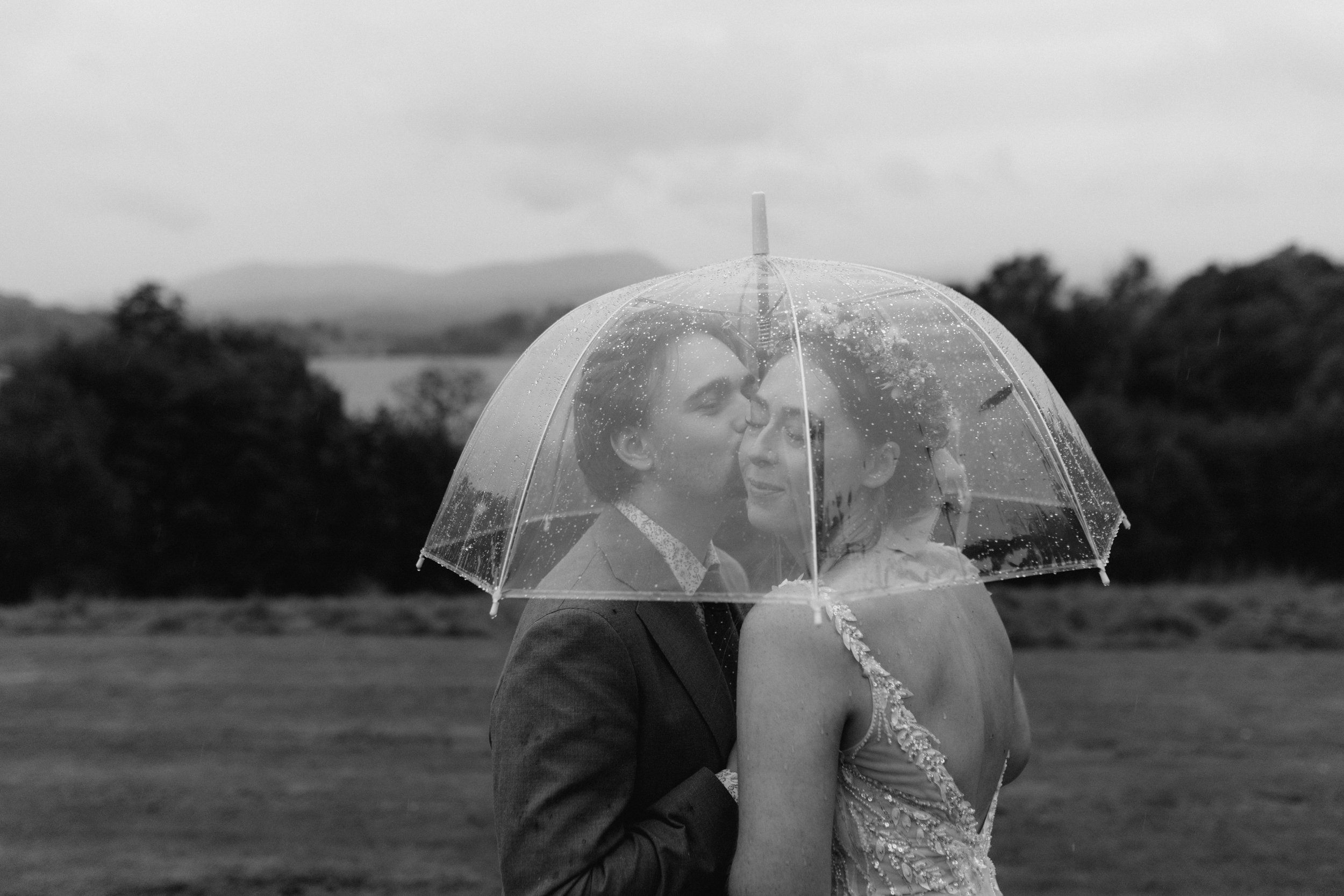  The groom kisses the brides cheek under an umbrella during a rainy Lake District wedding. 