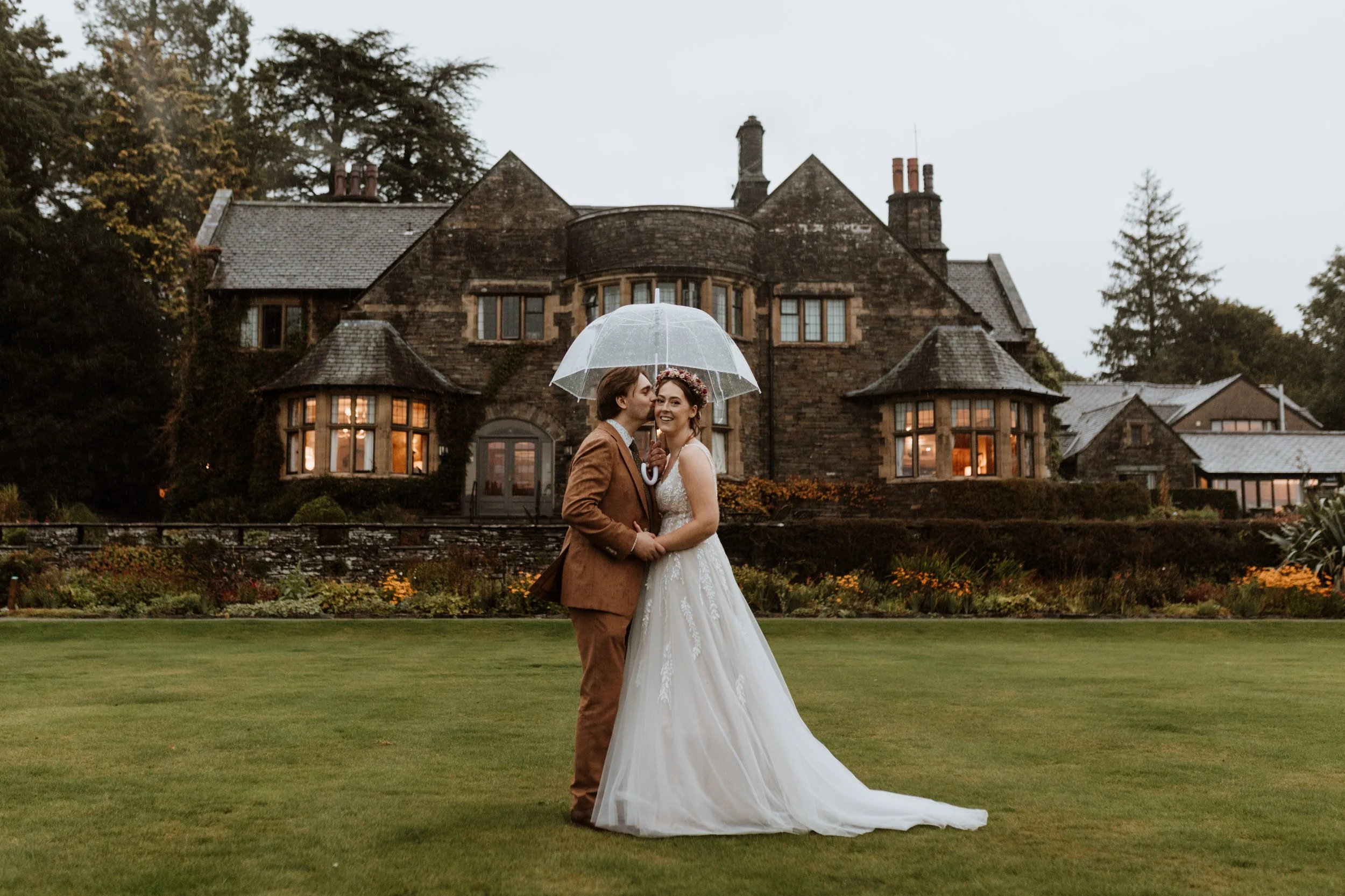  Bride and groom under an umbrella during a rainy Lake District wedding, with Cragwood Hotel in the background.  