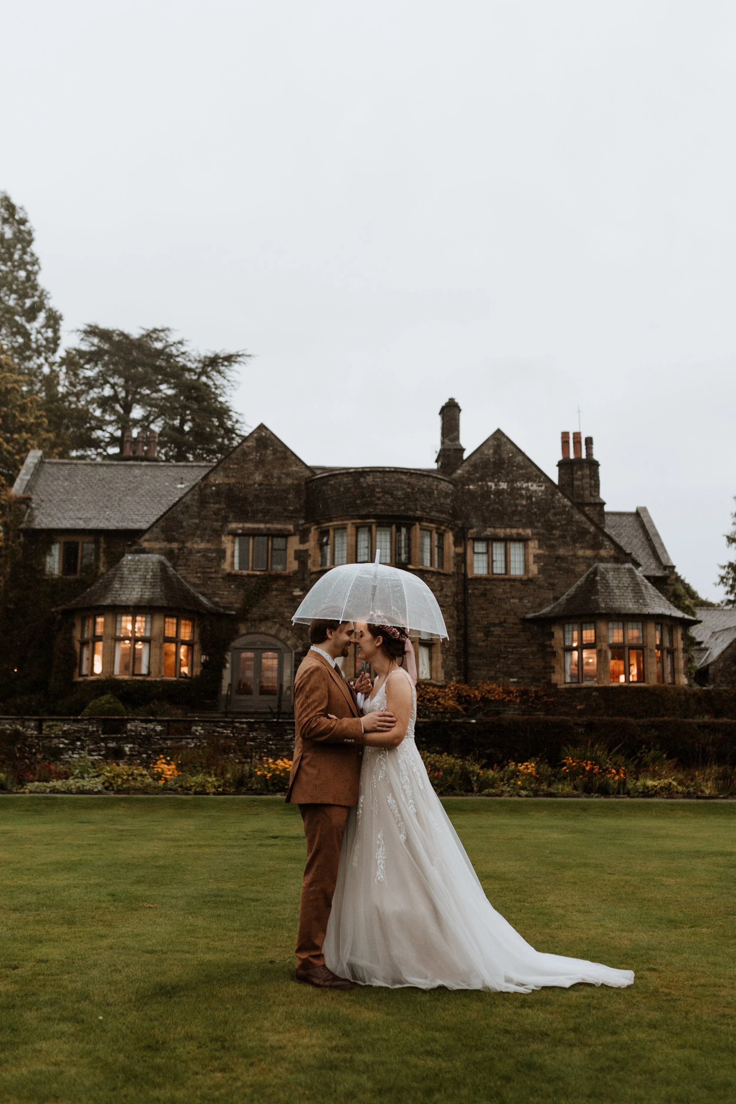  With Cragwood hotel in the background, the bride and groom stand under an umbrella during a rainy Lake District wedding 