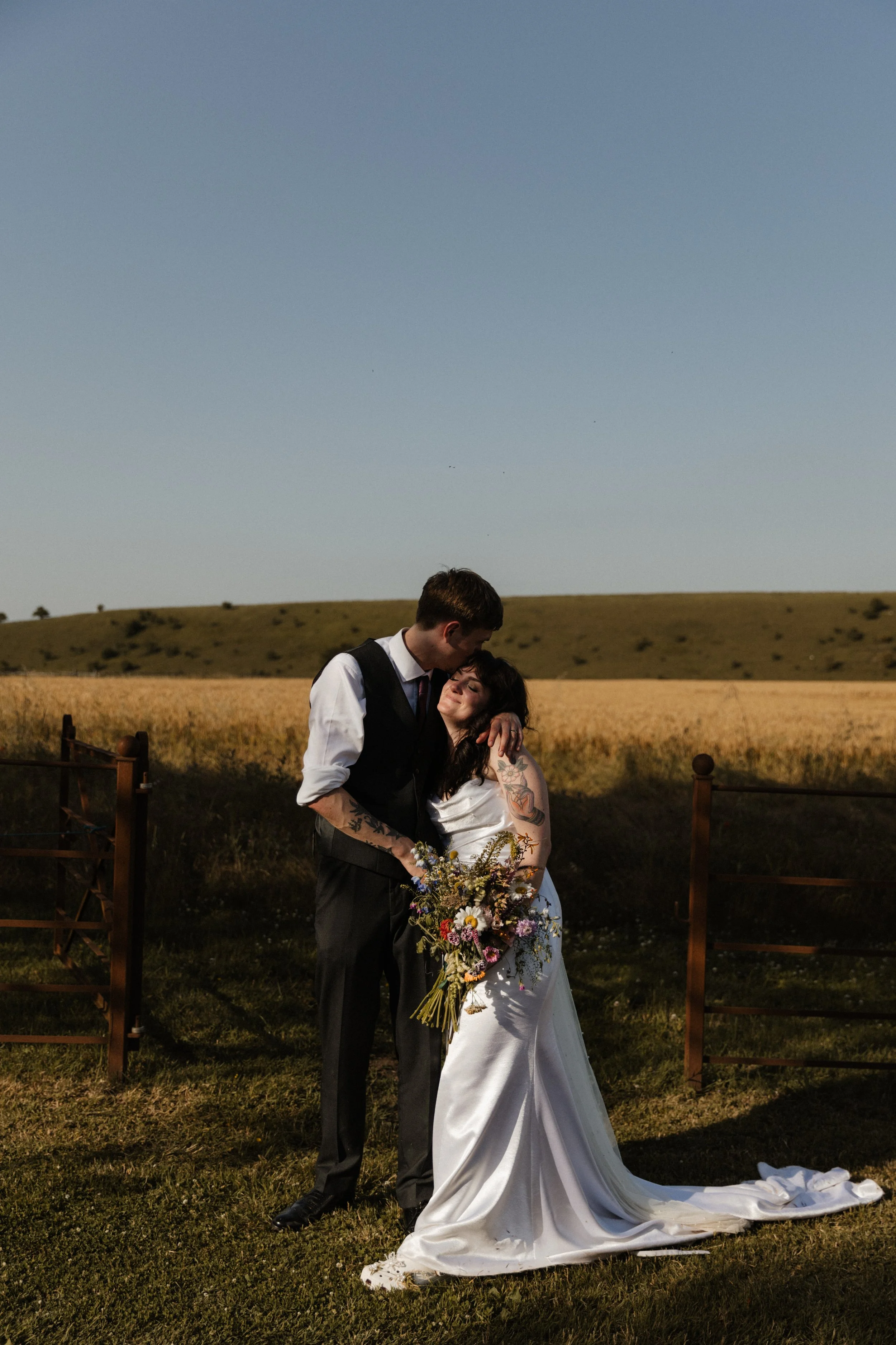 A groom kisses his bride on the forehead as they stand in front of a field of wheat.