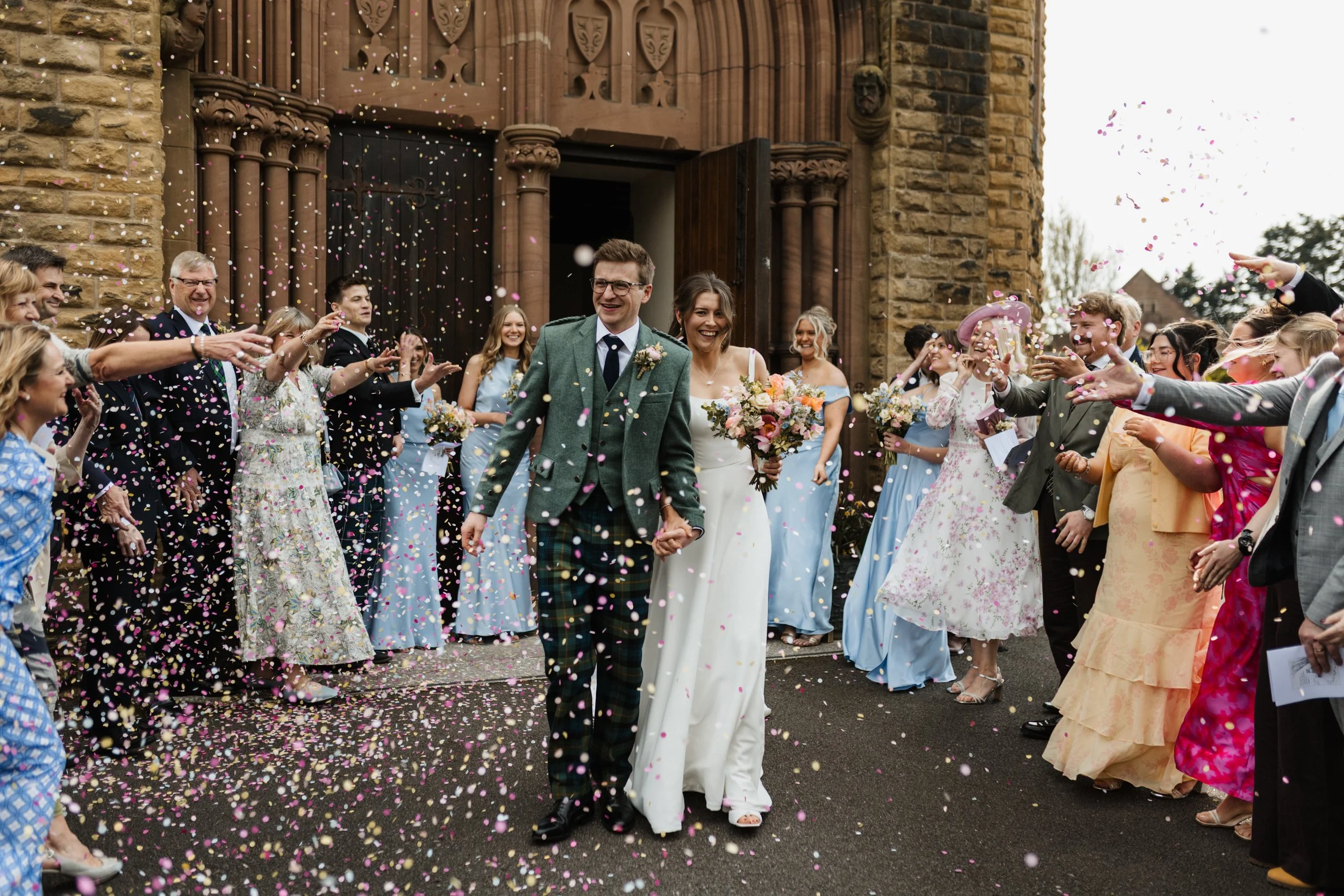 A bride and groom walk through a shower of confetti outside a church.