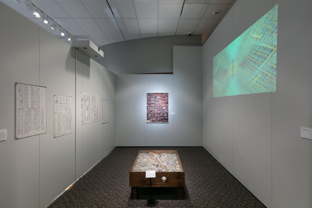 Room featuring enlarges project notes on the left wall, "Yosemite Table" in the fore gound, "Sand Hills 1" in the background, and a projection of multiple video projects on the right wall