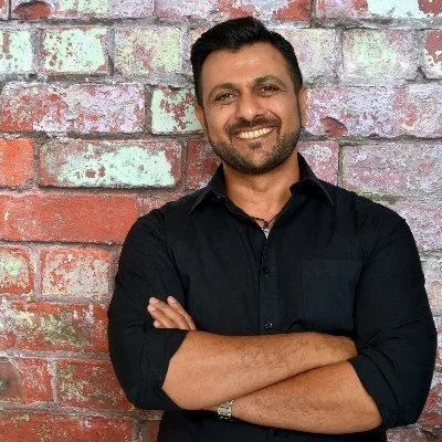 This is an image of Kul Mahay in front of a brcik wall with his arms crossed, and a big smile.