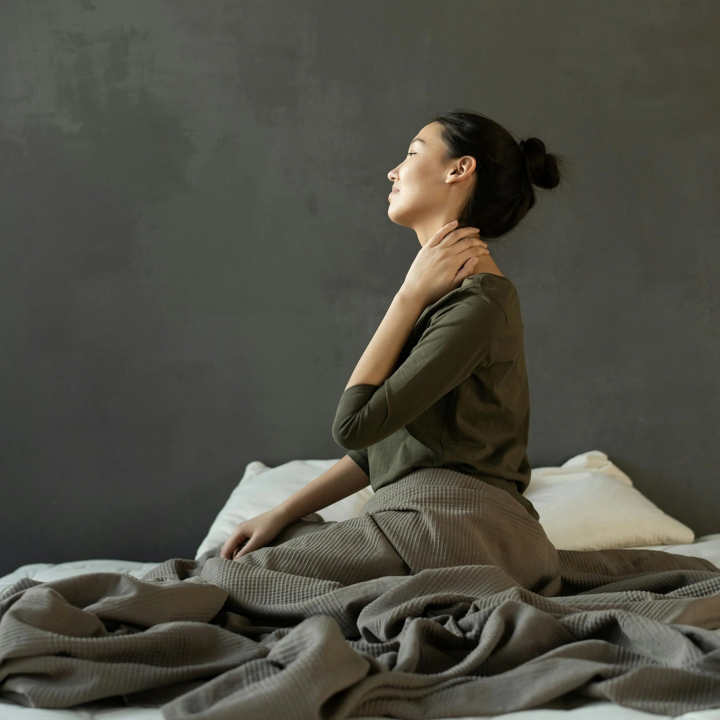 Woman sitting on a bed, holding her neck from pain in a dim room