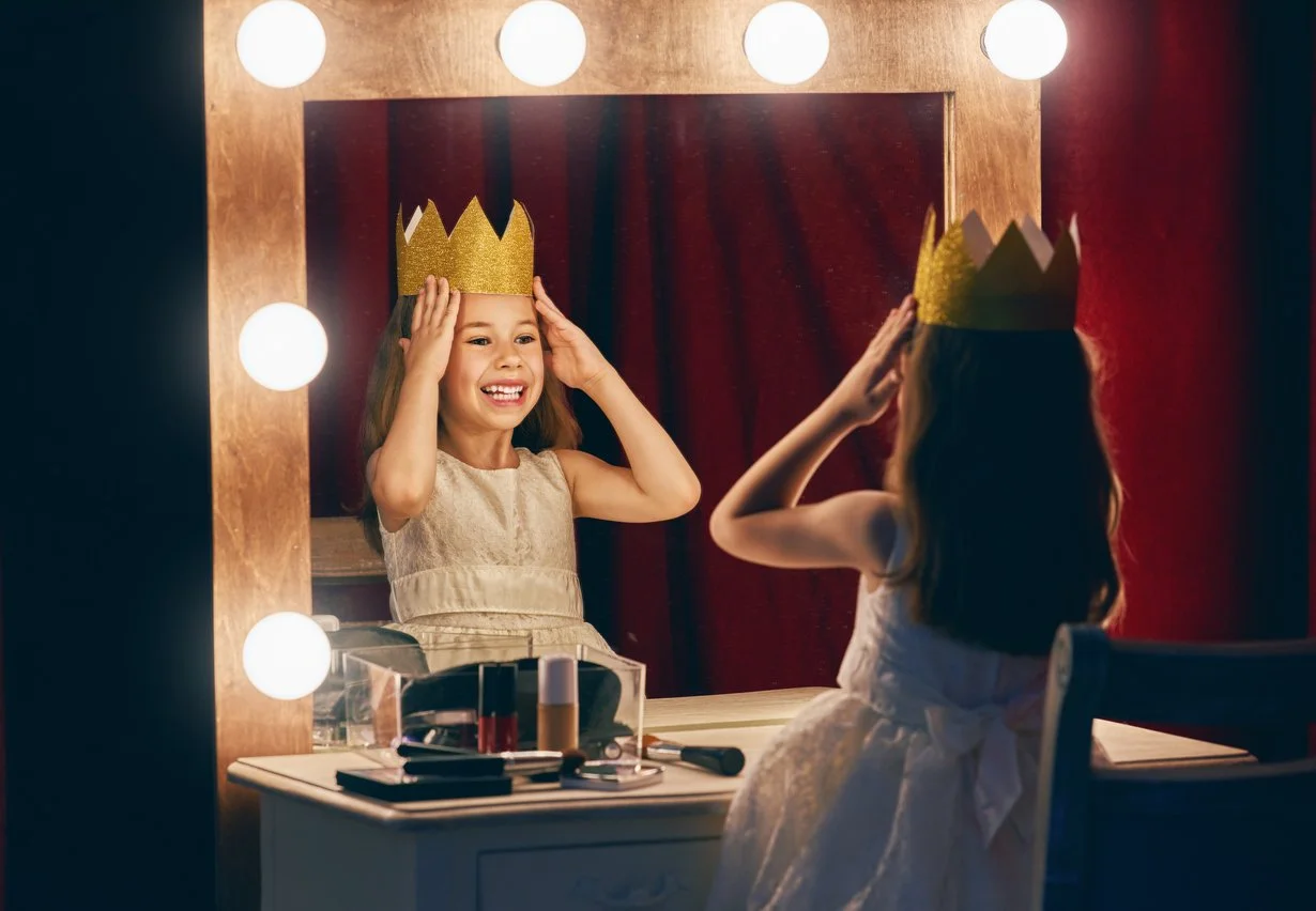 Girl in a princess costume putting on a gold crown in front of a mirror with a red theater curtain