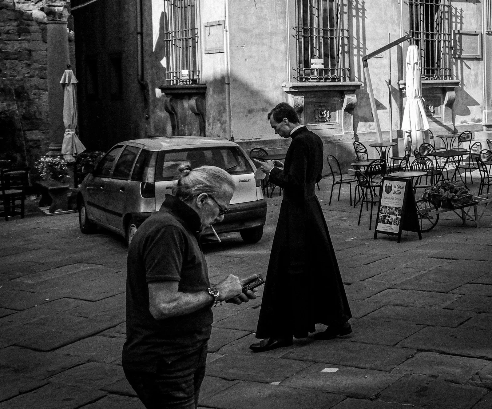 Two men are in a city street, one is standing and looking at a phone, wearing a long skirt and jacket, the other is an older man smoking and looking at a phone, wearing glasses and a watch.