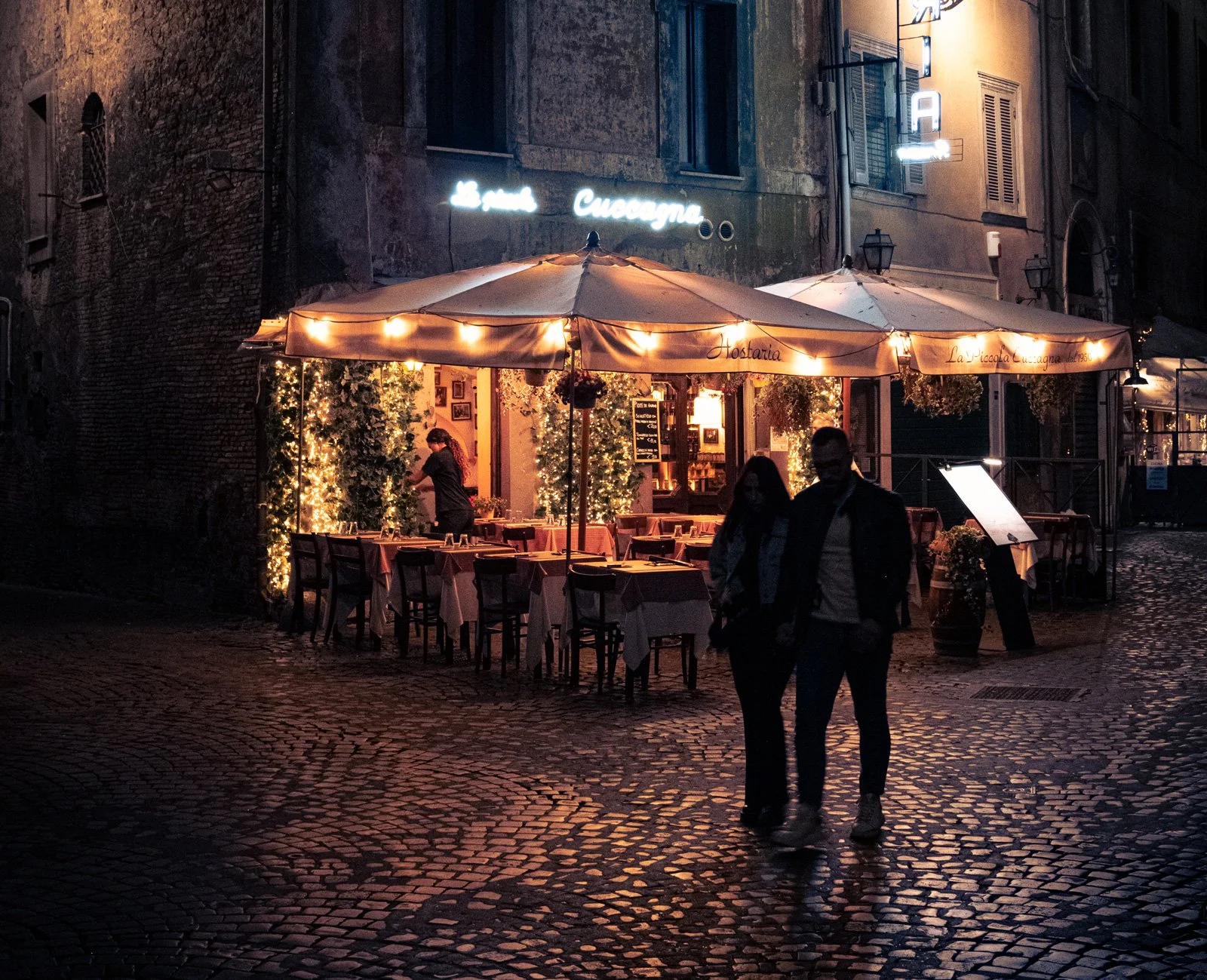 A couple walking on a cobblestone street at night in Rome in front of a warmly lit outdoor restaurant with tables, chairs, and string lights under a large beige umbrella, with buildings and signs in the background.