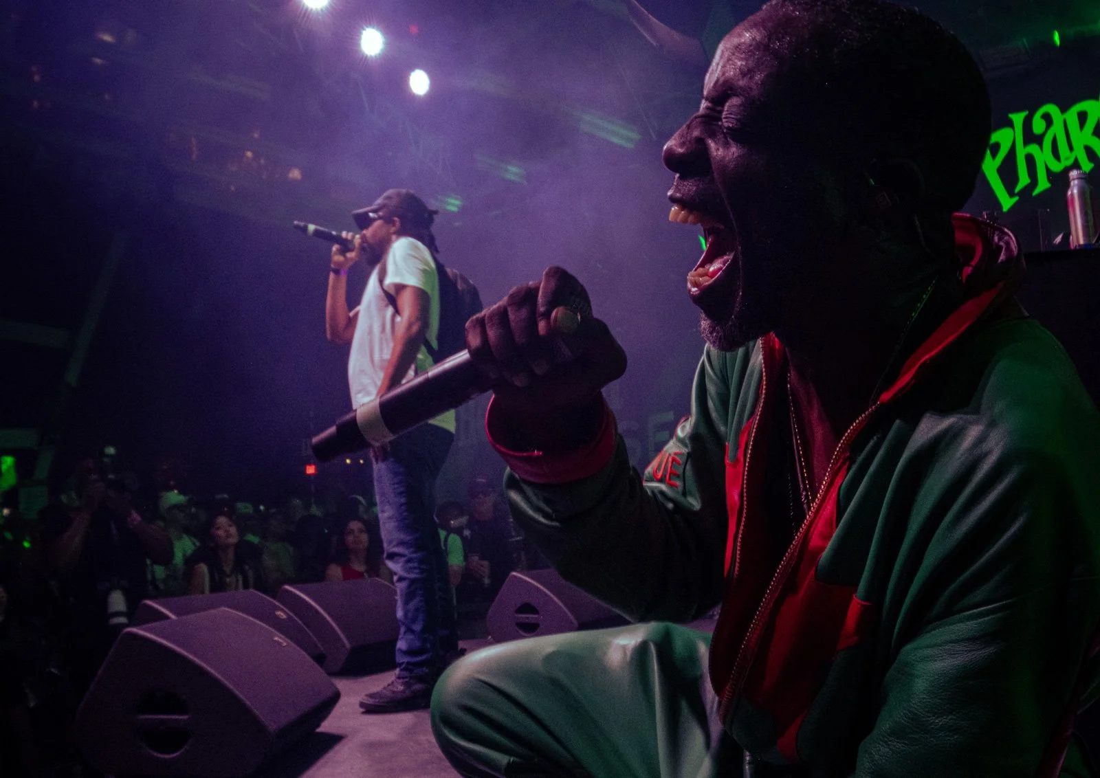 The Pharcyde singing into microphones on stage in a dark, colorful concert venue with an audience.