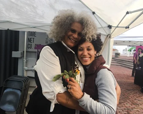 Lolita Parker, Jr. and Elisa Hamilton embrace after a slide talk; Elisa clutches garden seeds gifted to her by Lolita. Photo courtesy of the artist.
