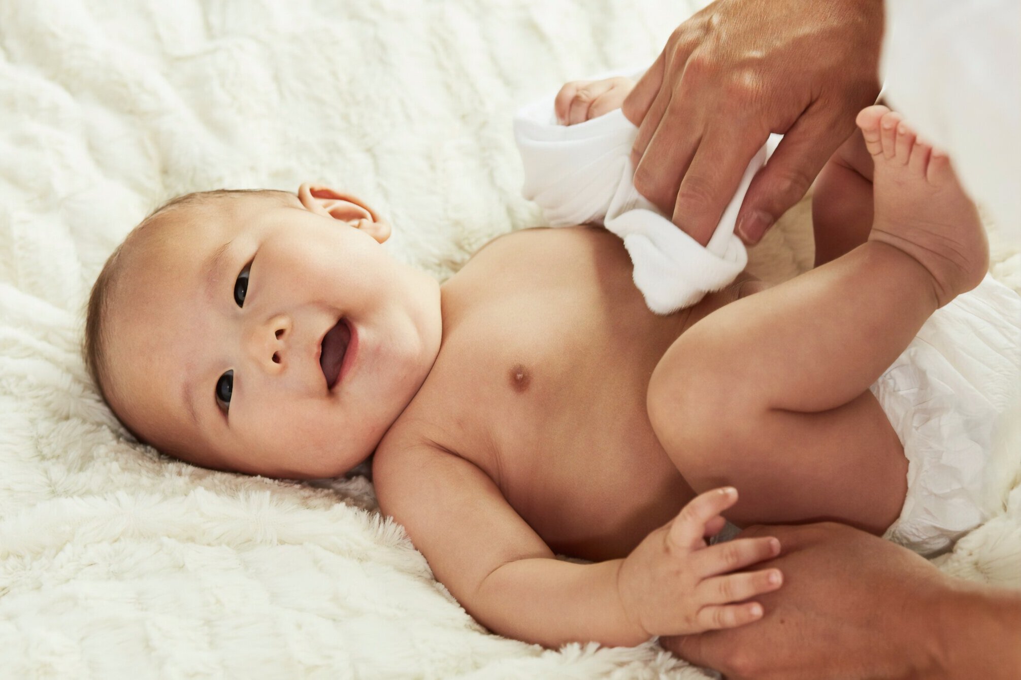 Img Description: Smiling Baby in diaper lying on cozy blanket, parent’s hand holding a cloth and wiping his belly