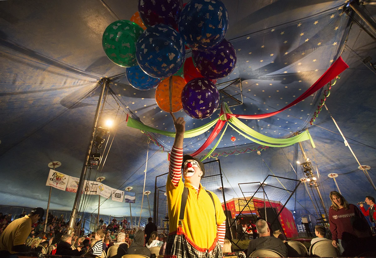 John Sayre, aka J.P. Ballyhoo, balances balloons on his finger while vendors sell peanuts to the crowd and the crew prepares the arena for the next act.