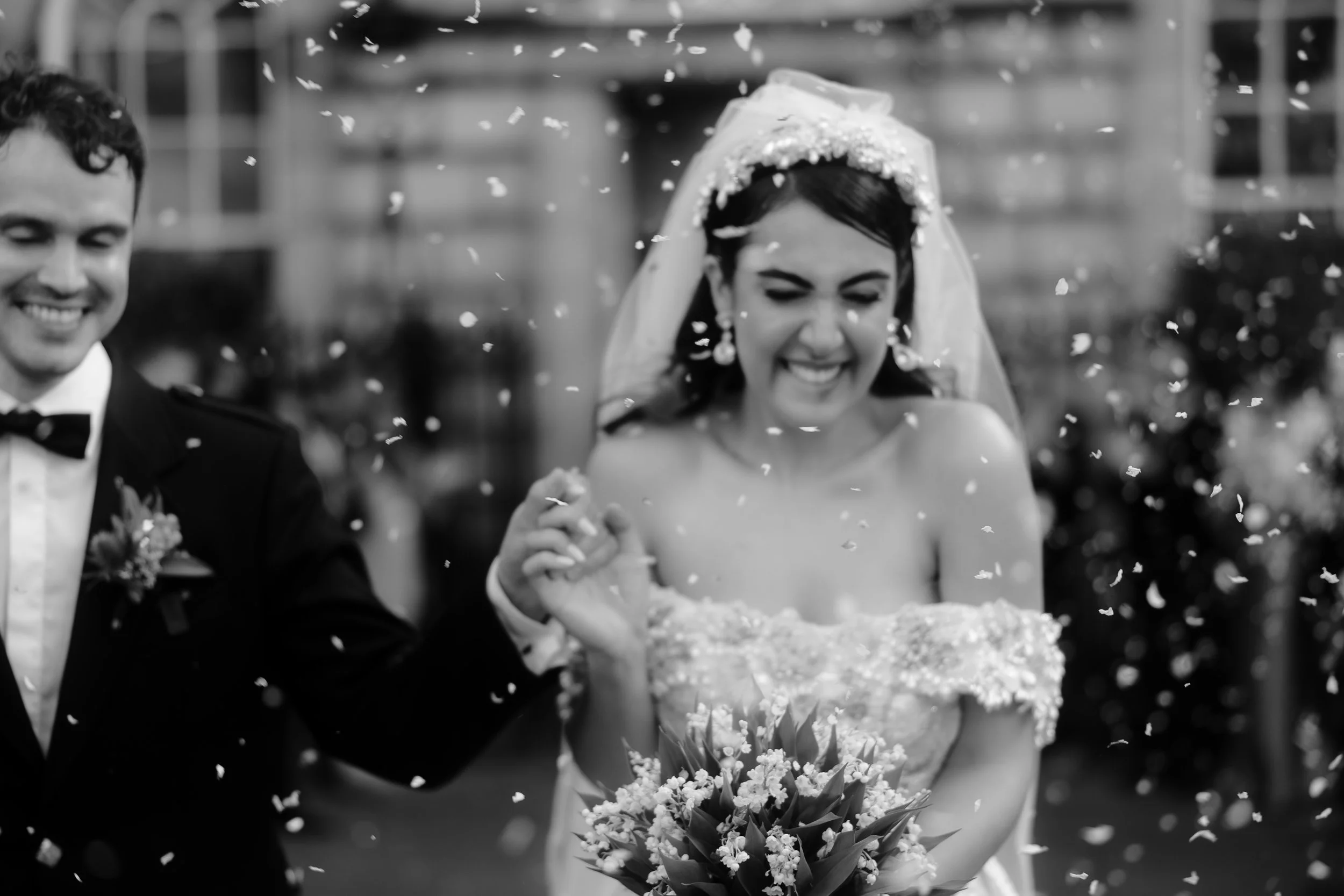 Black and white photo of a bride and groom smiling and holding hands during their wedding celebration, with confetti falling around them.