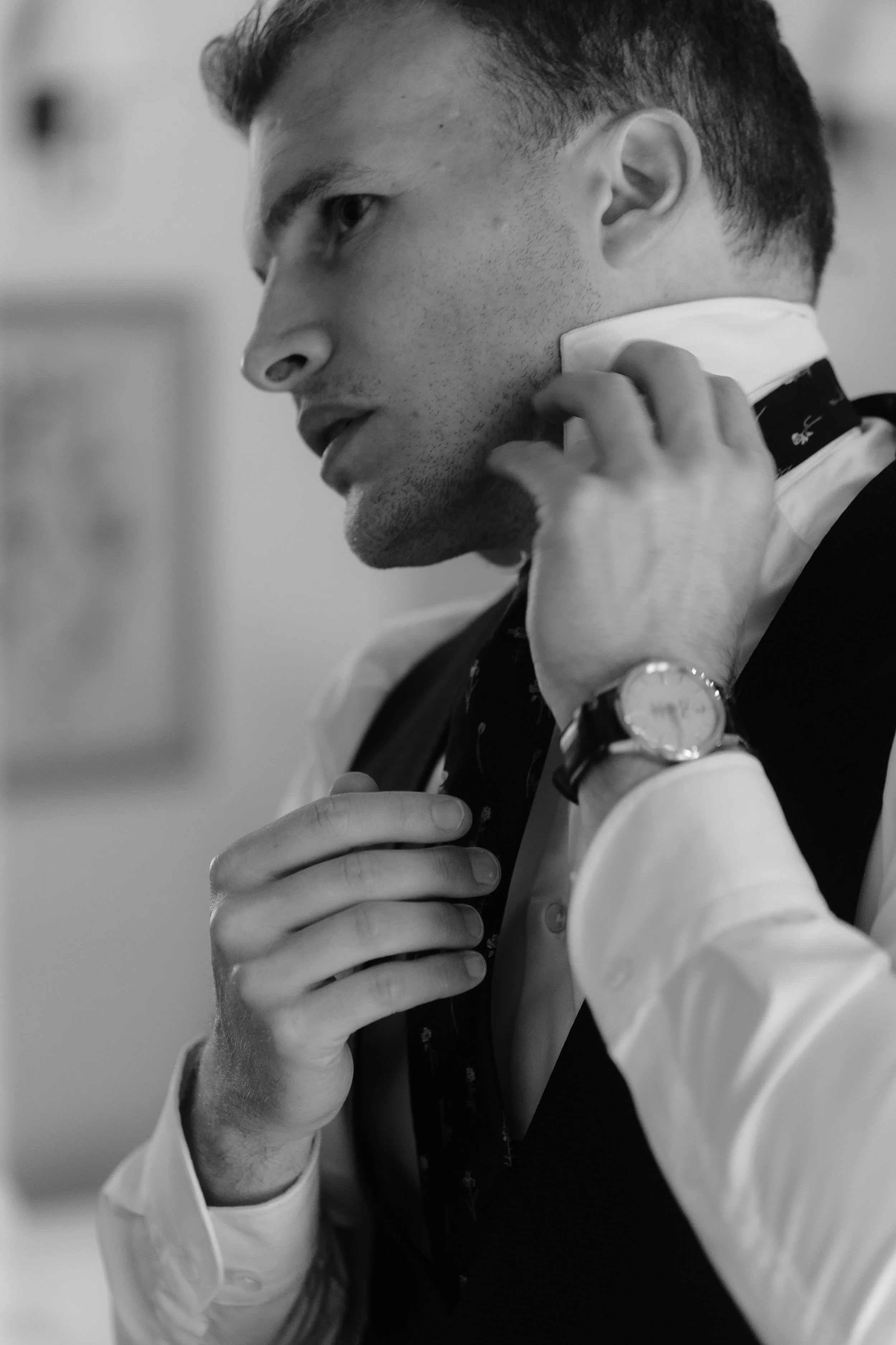 Black and white photo of a man adjusting his collar, wearing a watch, a shirt, vest, and tie, with a focused expression.