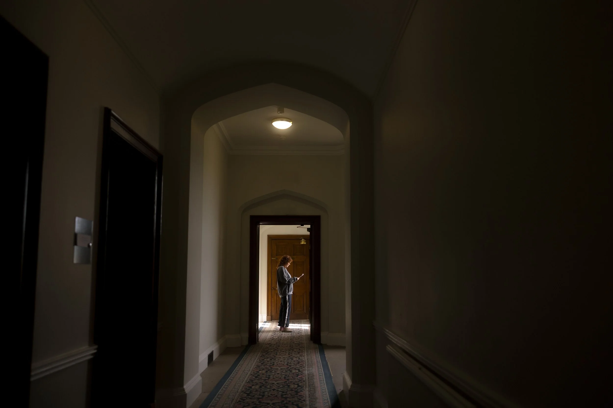 Person standing in a hallway at a doorway, looking at a mobile device, illuminated by natural light from outside.