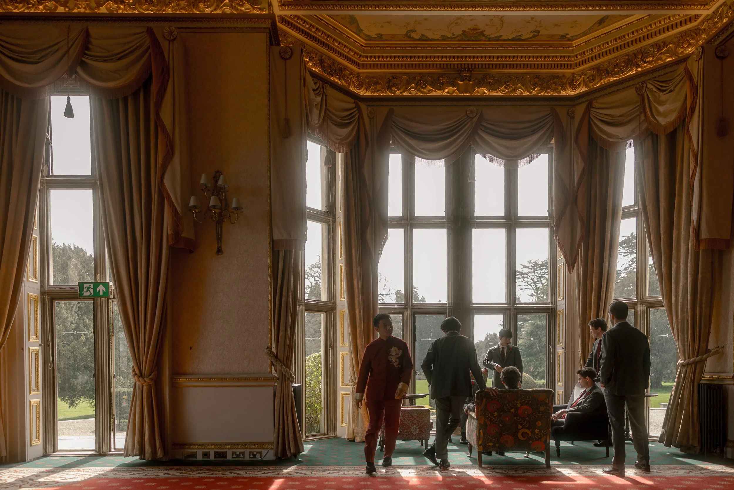 Group of people in a luxurious room with tall windows, draped curtains, and ornate ceiling, engaged in conversation and seated on chairs.