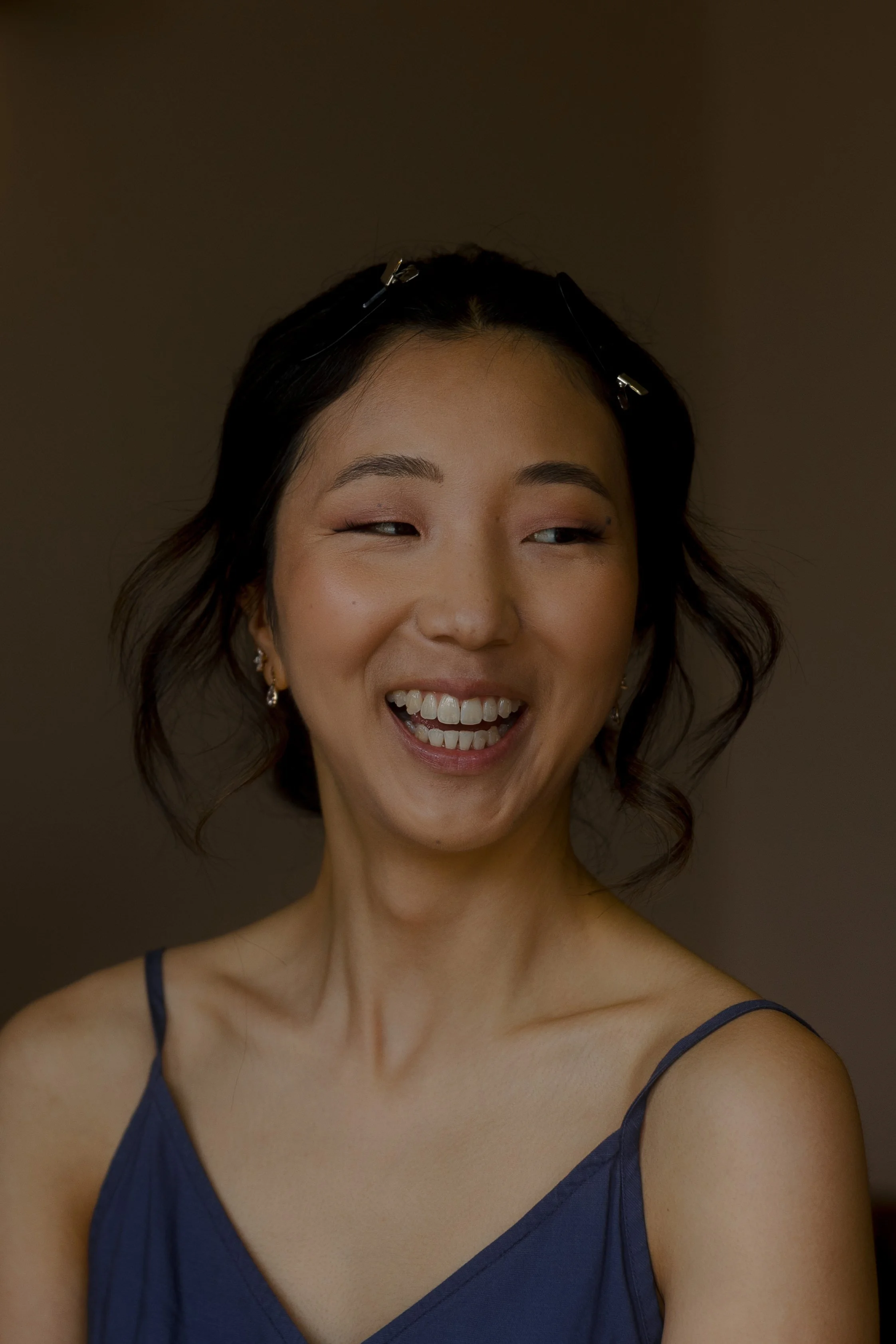 Close-up of a smiling East Asian bride with wavy black hair, wearing a blue spaghetti strap top and earrings, looking to the side.