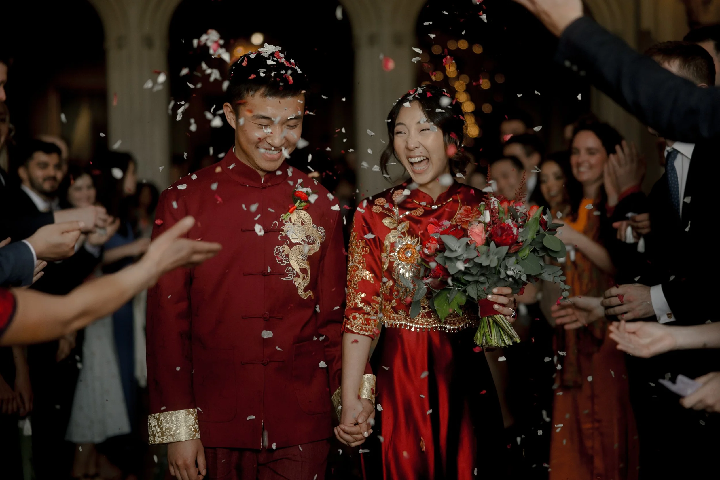 A couple dressed in traditional red Asian wedding attire holding hands and smiling, surrounded by guests throwing white and pink confetti during a wedding celebration.