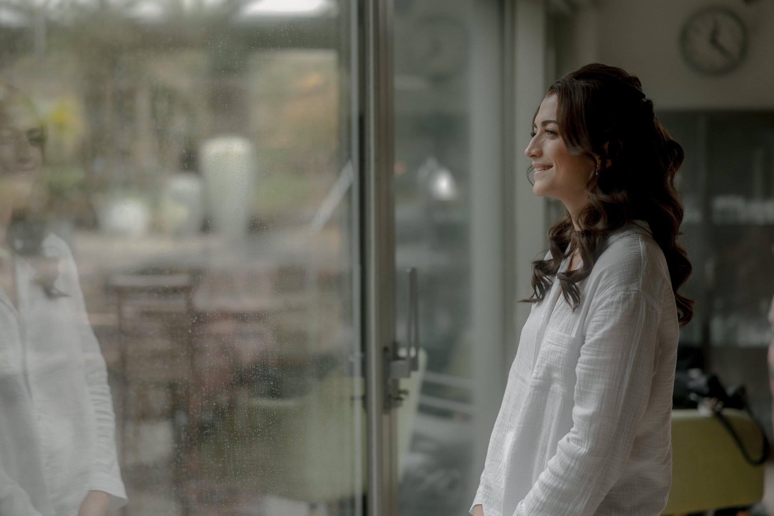 A woman with long dark curly hair, wearing a white shirt, stands indoors smiling and looking out a rainy glass door, with her reflection visible on the wet glass.