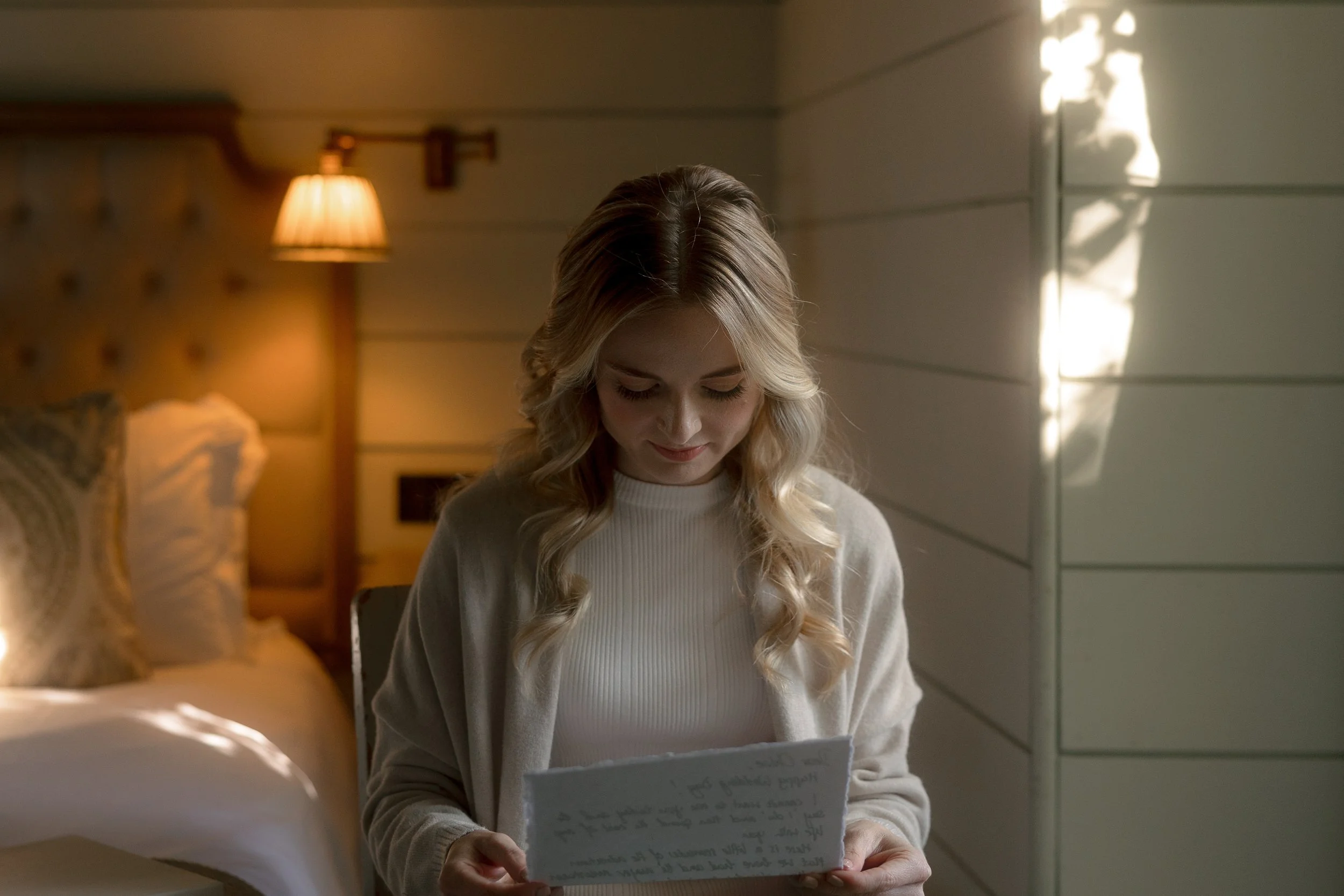 A young woman with blonde hair reading handwritten letters in a warmly-lit bedroom.