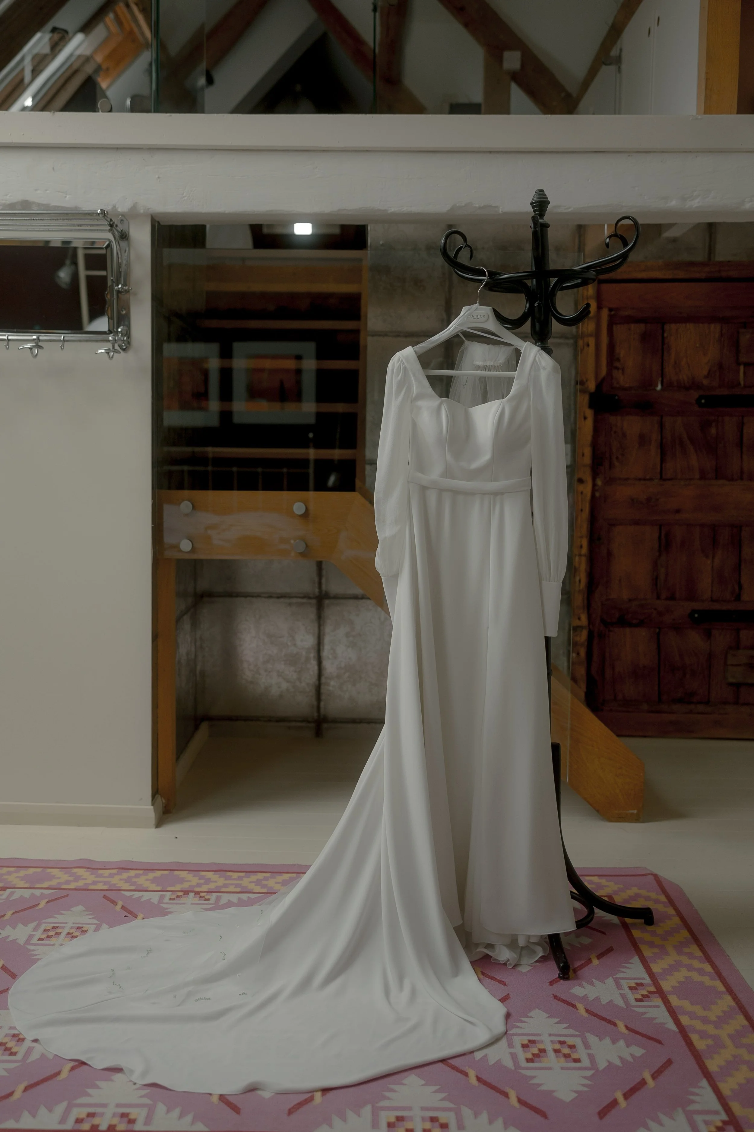 A white wedding dress hanging on a coat rack in a room with wooden walls and a pink patterned rug.