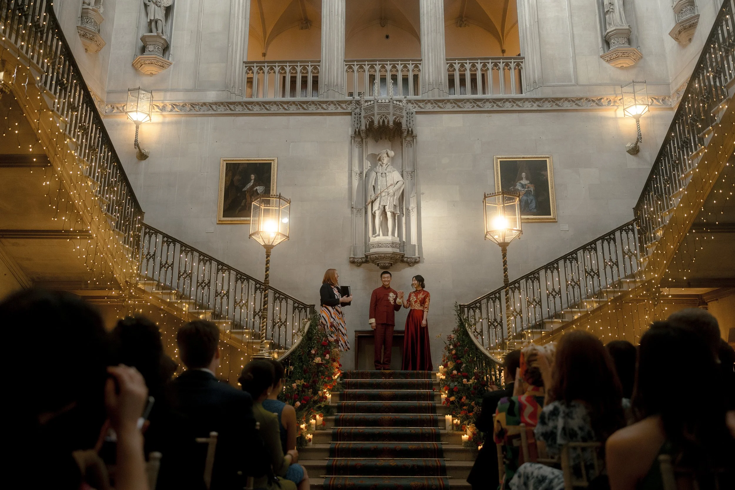A wedding ceremony taking place on a grand staircase inside a historic building with Gothic architecture. The bride and groom are standing at the top of the stairs with an officiant, surrounded by guests. The staircase is decorated with flowers and c