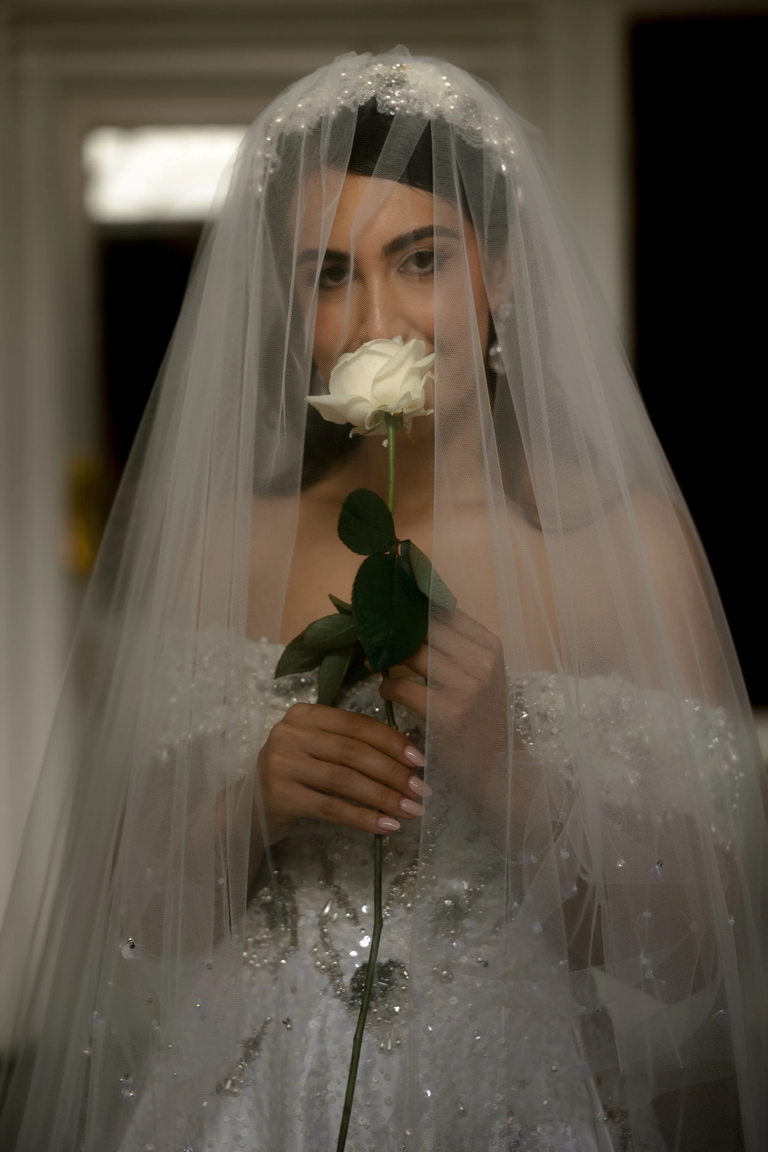A bride in a wedding dress and veil holds a white rose in front of her face, partially obscuring her smile, standing indoors.