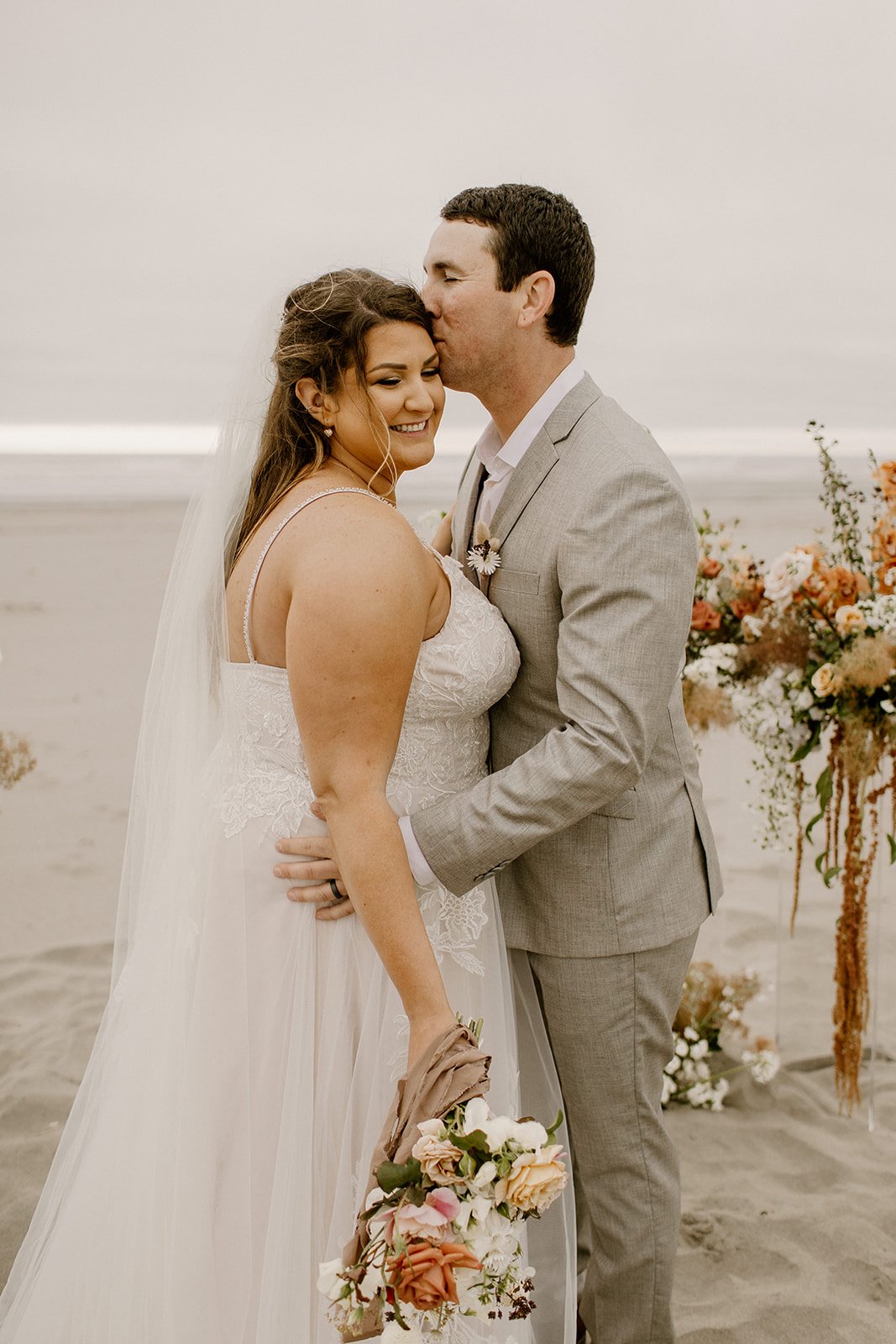 groom kissing bride on the temple as she looks down at her bouquet while standing on the beach in coastal washington