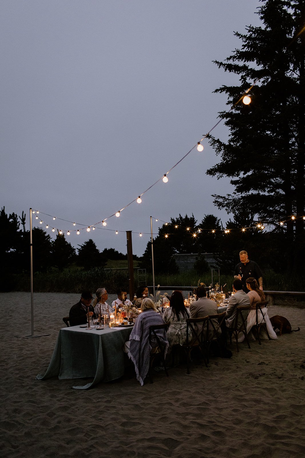 night time photo of guests sitting outside on the beach enjoying dinner and toasts 