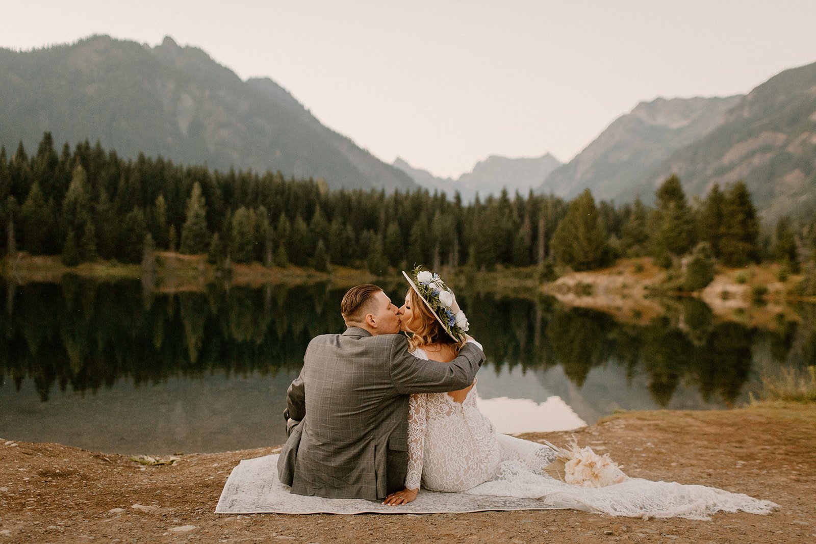 couple sitting on a blanket on the edge of a lake while kissing in their wedding attire in washington