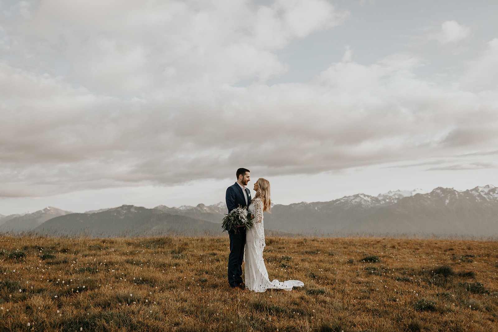 Bride and Groom in their wedding attire standing in a meadow with mountains in the backdrop