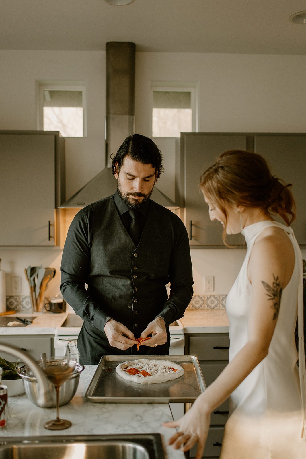 Groom in his wedding attire putting pepperoni on a pizza in the kitchen of their Airbnb while the bride watches in her wedding dress