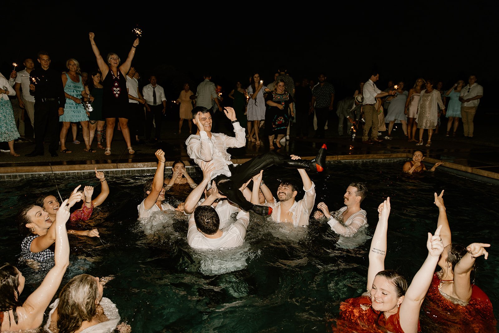 groom being lifted up in the pool by friends after he just jumped in with all his clothes on after his sparkler exit