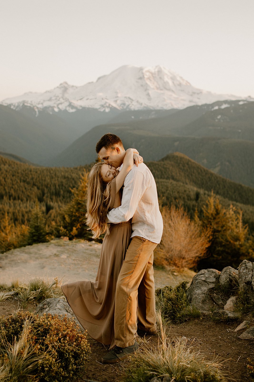 Women hugging her partner with her arms wrapped up around his neck while he is kissing her on the cheek while standing in front of mt. Rainier's peak