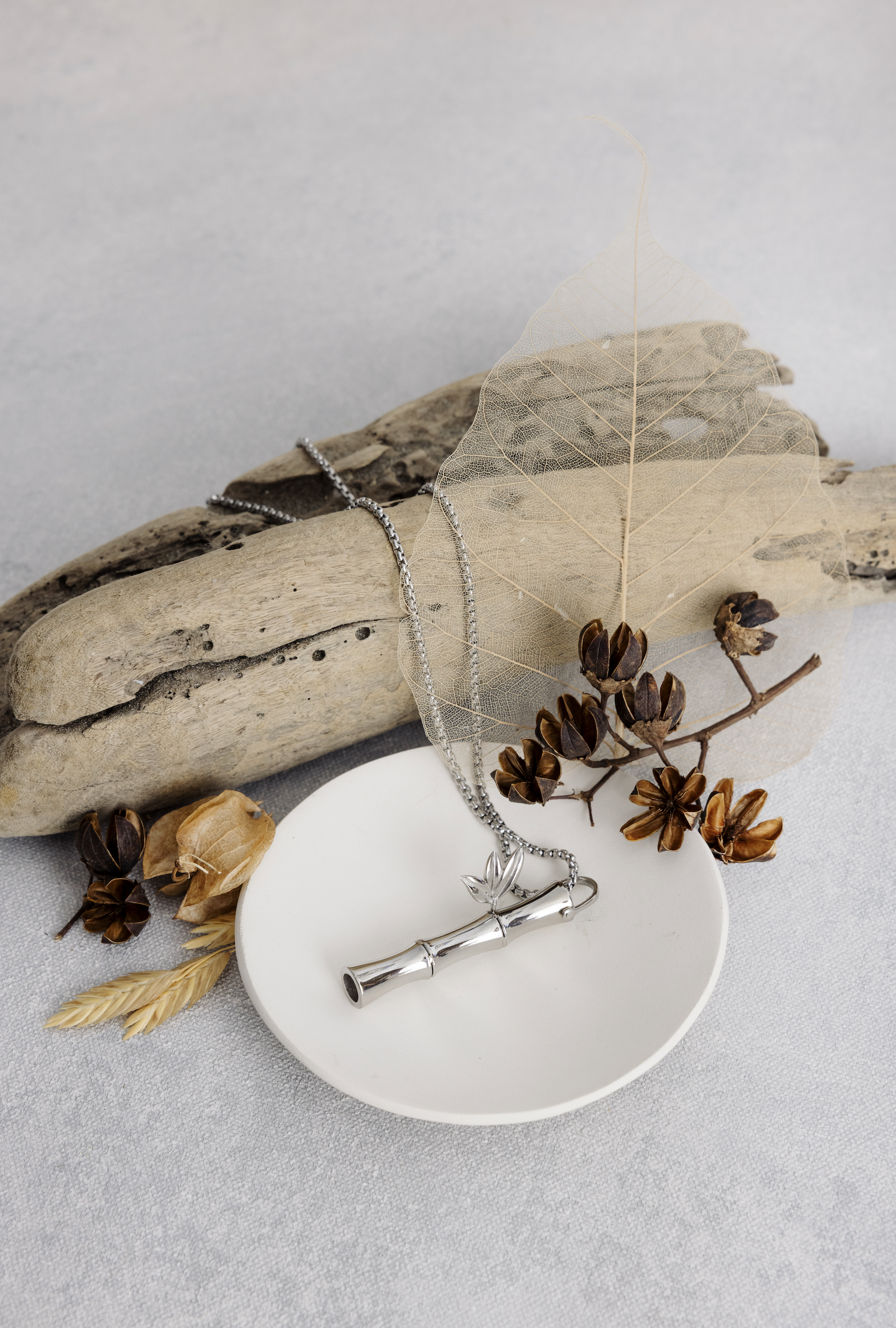 Silver necklace with leaf pendant on white ceramic dish, surrounded by dried leaves, seed pods, and a leaf skeleton on a gray fabric background. Shot for TOWN Magazine in Greenville, SC by Melissa Glynn