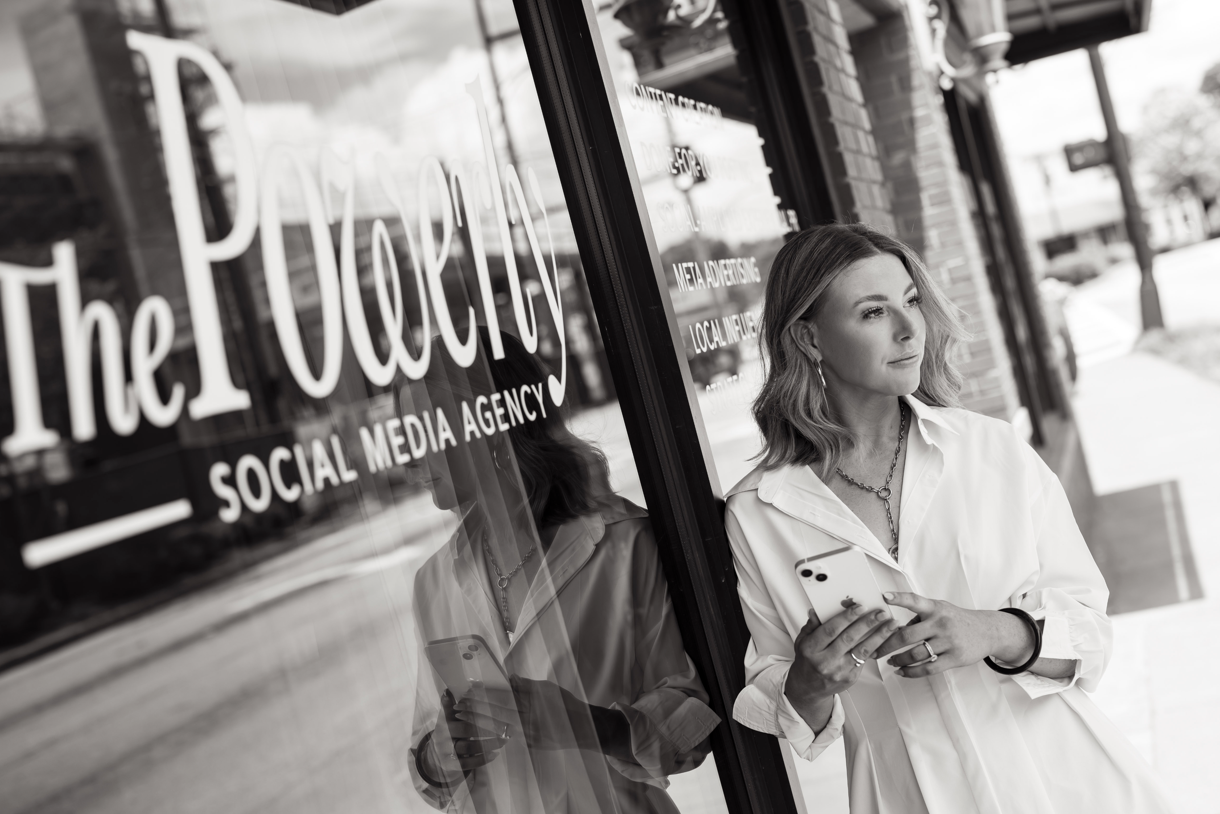 A woman with wavy hair wearing a white shirt, standing outside a business storefront, looking at her phone, with store signage reflected in the window. The Powerly Social Media Agency in Greenville, SC - Photo by Melissa Glynn
