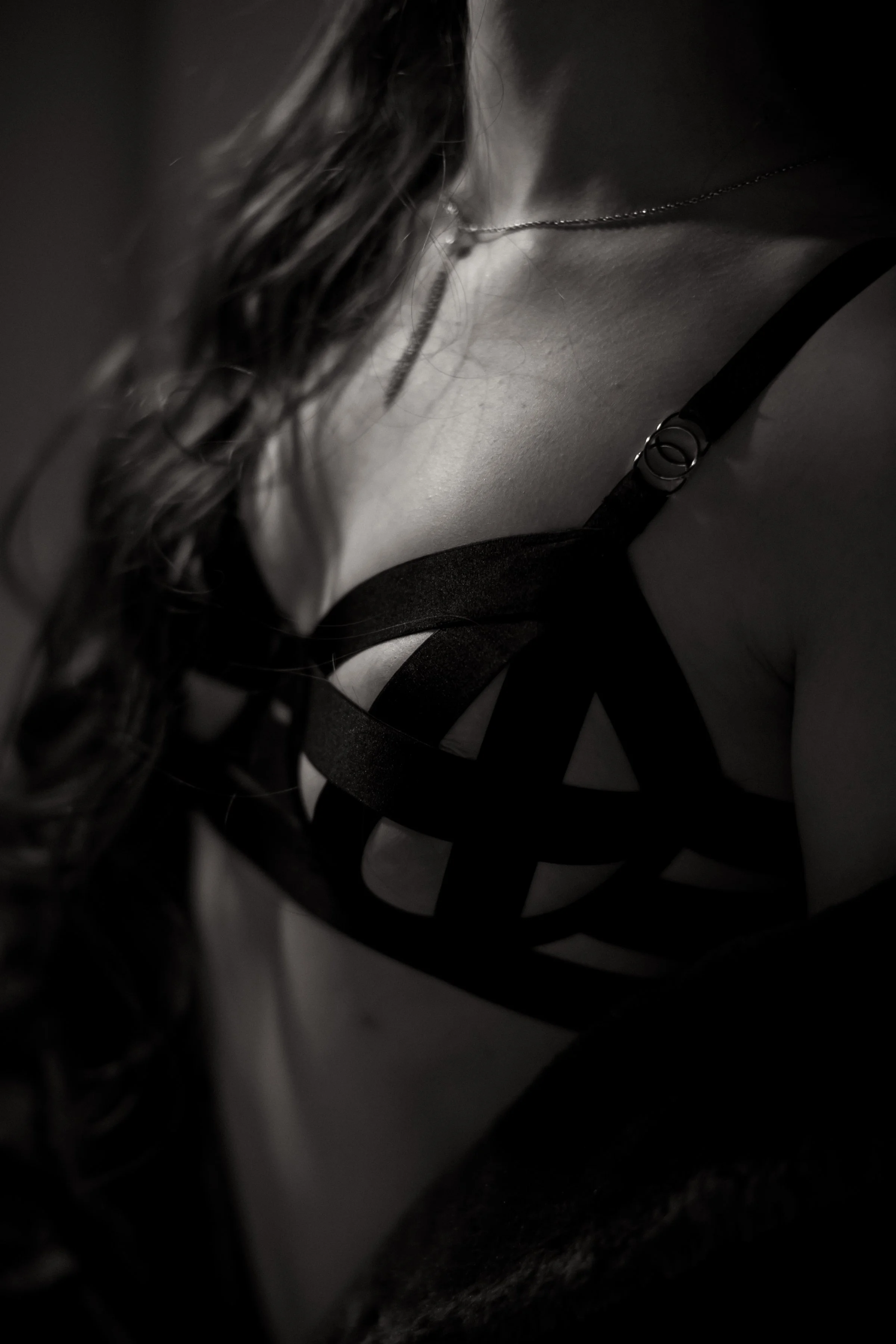 Close-up of a woman's chest and shoulder, wearing a black strappy top and a necklace, with long wavy hair.