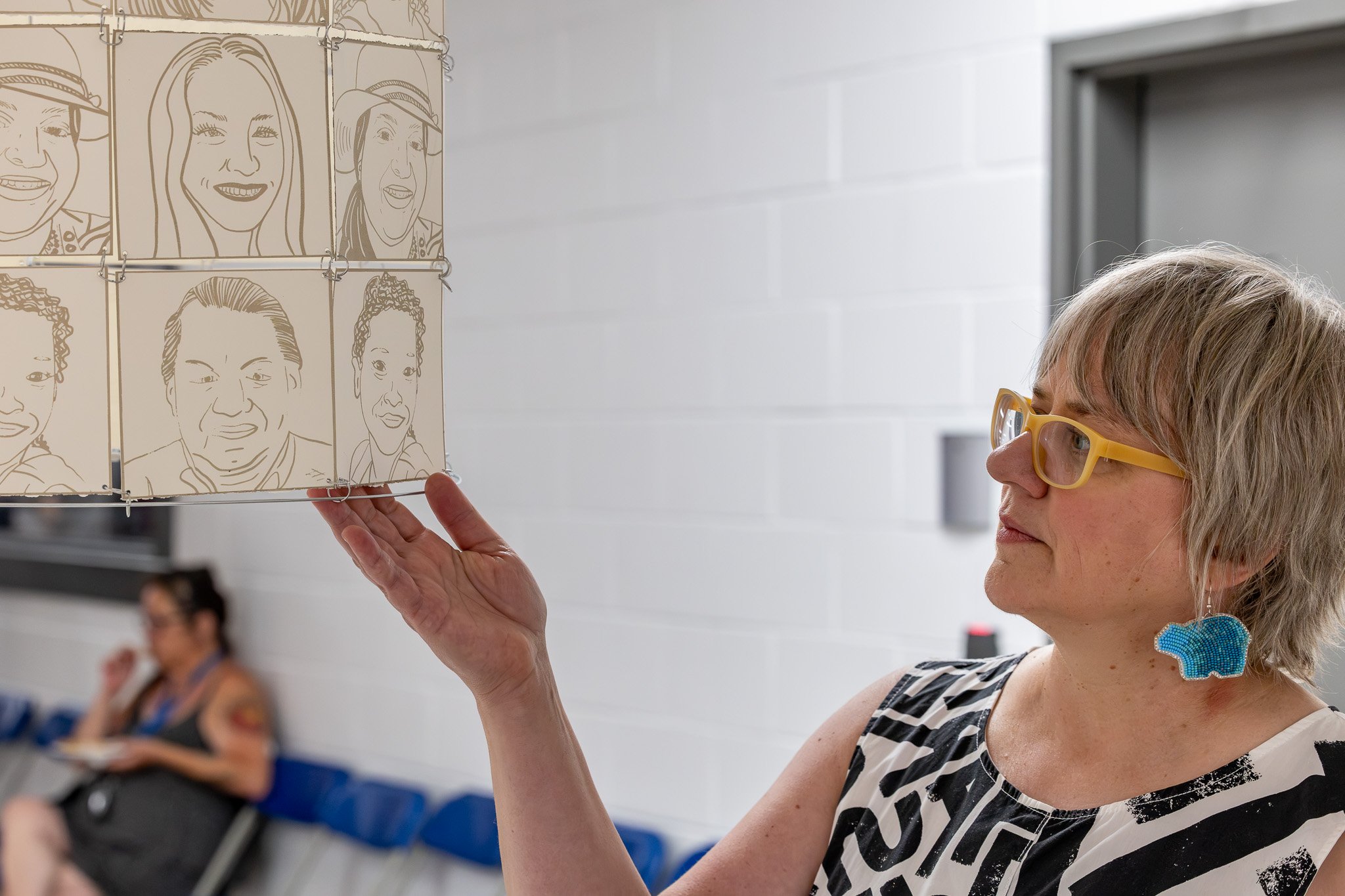 Pale-skinned woman with yellow glasses and blue bear earrings in the right side of the image holds the bottom of a paper-and-wire cylinder that shows portraits of several faces that are screen printed on the paper.