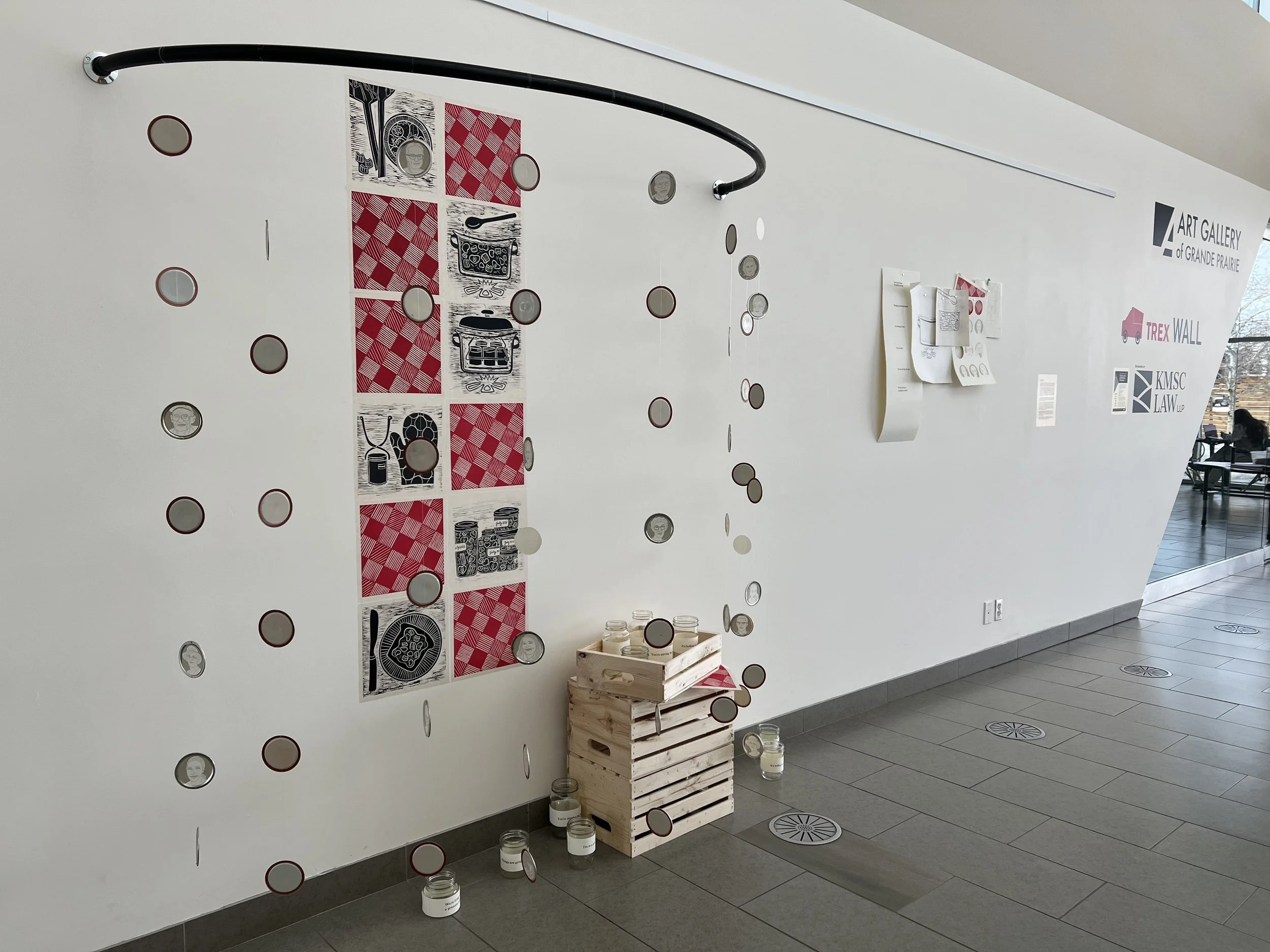 lino cut prints that tell the story of making a jar of jam lie on w wall behind a semi circle of hanging circles. The circles are screen printed portraits of community members attached to canning jar lids. On the floor are wooden boxes with jars